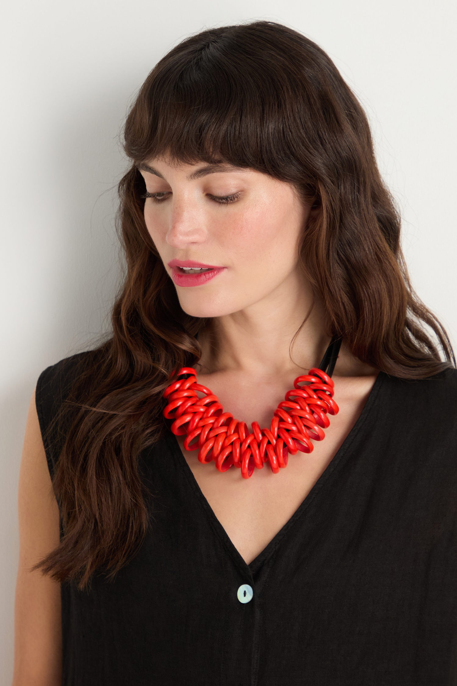 Woman with wavy brown hair wearing a black v-neck top and the Murano Links Necklace—a chunky red statement piece handmade in Paris—looking down slightly with a neutral expression.