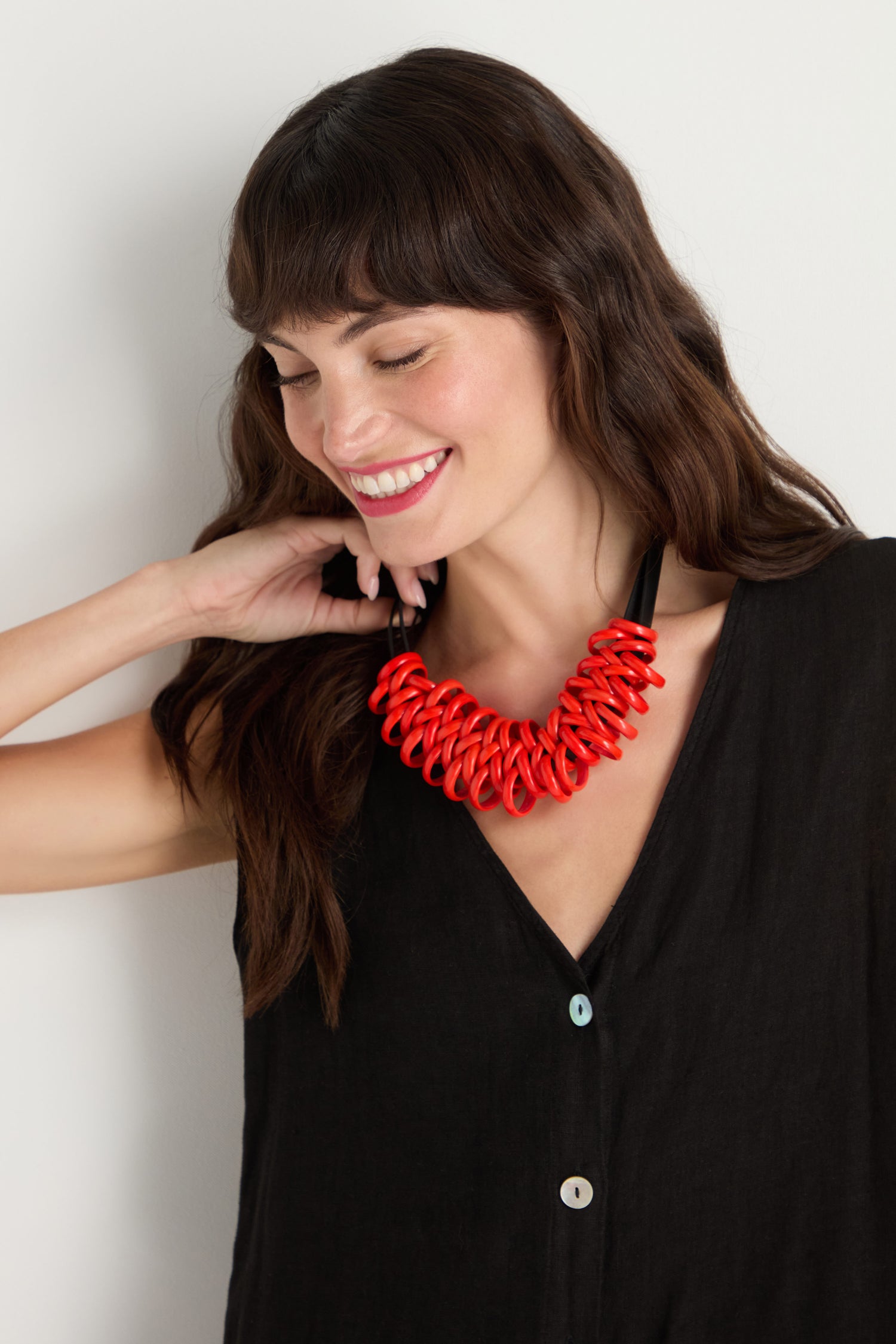 A woman with long brown hair smiles and looks down, wearing a sleeveless black top and the Murano Links Necklace—a bold red, handmade Parisian statement piece.