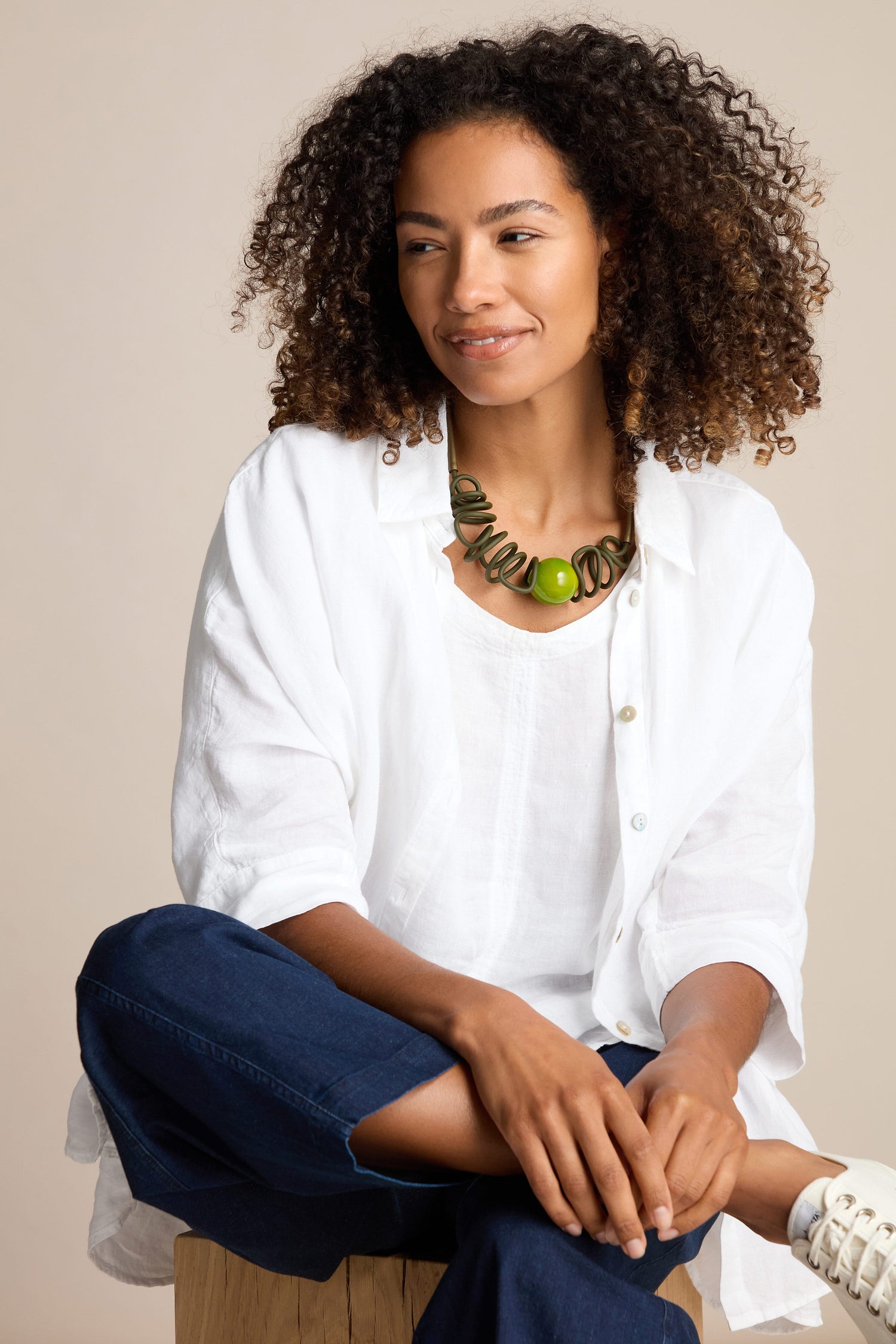 Person with curly hair wearing a white shirt, blue jeans, and a Samuel Coraux Murano Sketch Necklace, sitting with legs crossed on a wooden stool against a beige background.