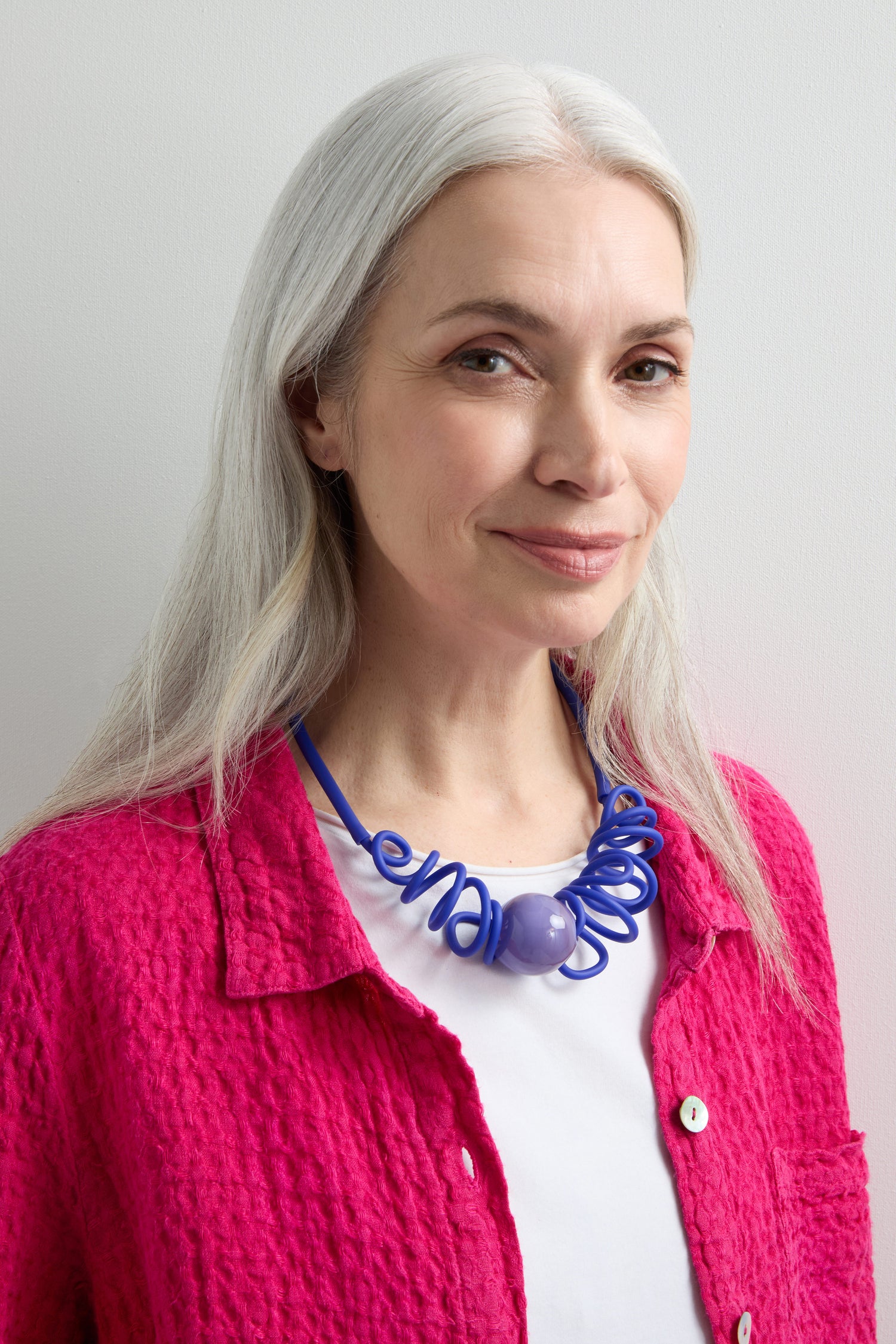 A woman with long white hair, dressed in a bright pink jacket and white shirt, is smiling slightly while wearing the striking Murano Sketch Necklace against a plain background.