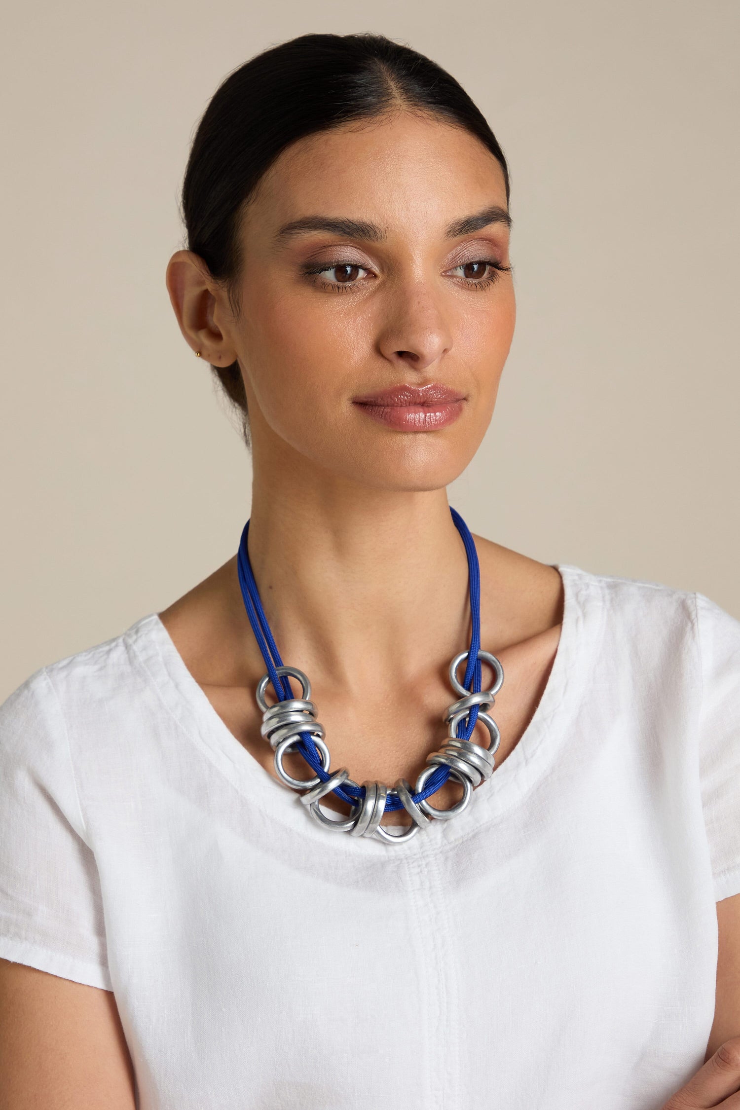 A woman wearing a white top and a Samuel Coraux Metal Rings Necklace, featuring large blue and silver rings. She has dark hair tied back and is looking slightly to the side against a plain background, showcasing her sculptural accessory.