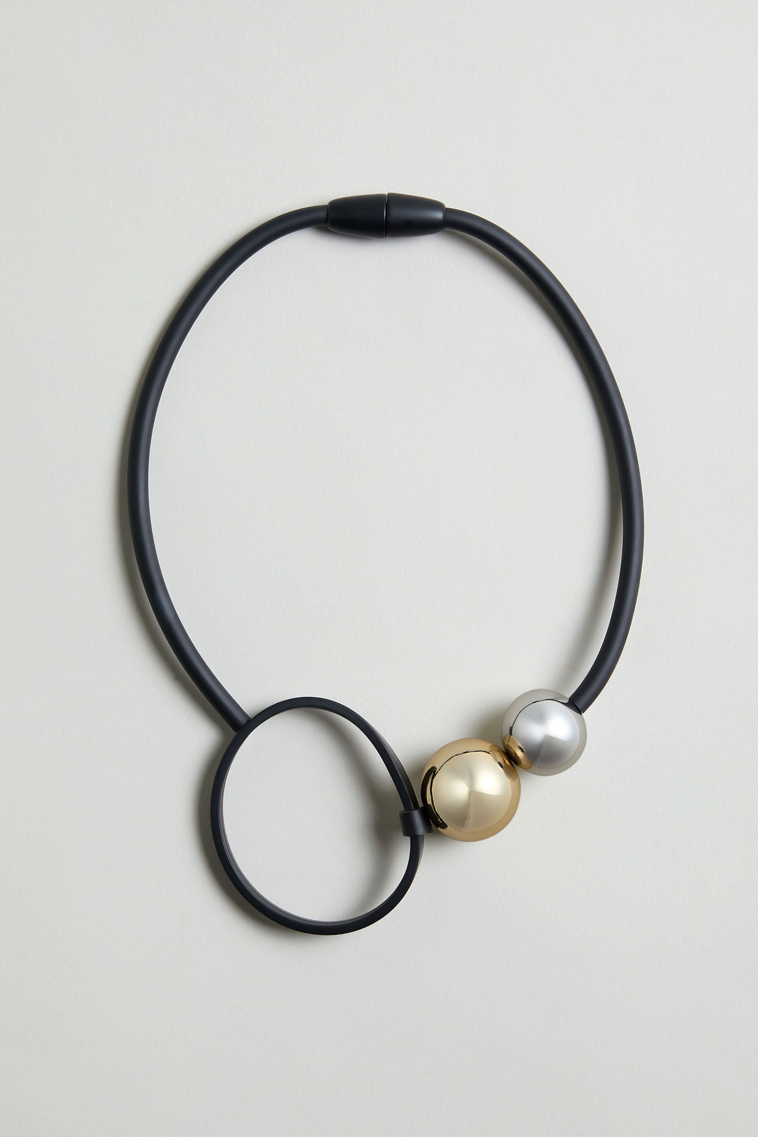 The Space Necklace features black rubber, a large circular loop, and two oversized acrylic beads—one gold, one silver—near the clasp.