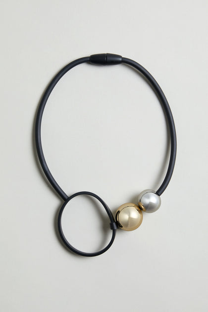 The Space Necklace features black rubber, a large circular loop, and two oversized acrylic beads—one gold, one silver—near the clasp.