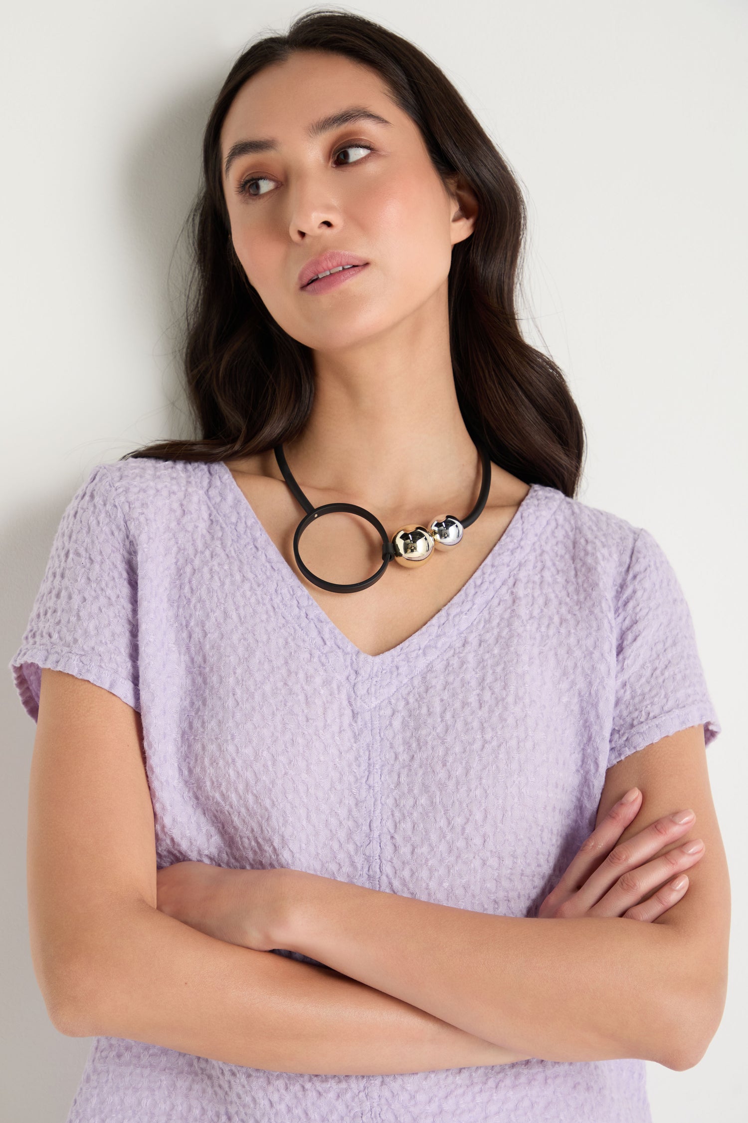 A woman with long dark hair, wearing a textured light purple top and the Space Necklace—an eye-catching piece with oversized acrylic beads—stands with arms crossed and gazes slightly to the side.