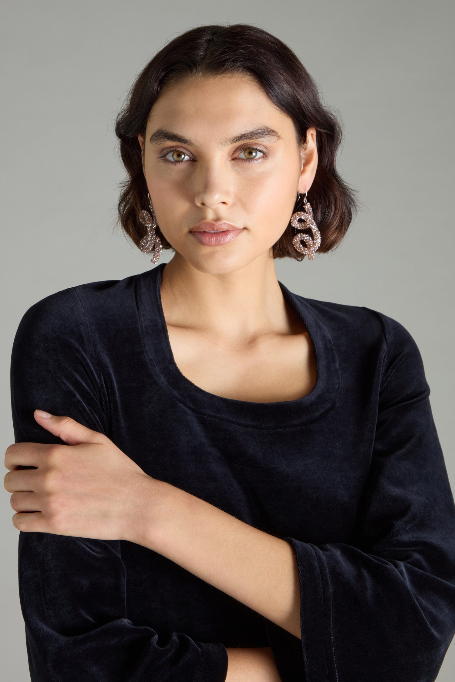 Sporting the Disco Sketch Earrings, an individual with short dark hair creates an artistic impression in a dark shirt, striking a pose with one arm crossed against a simple backdrop.