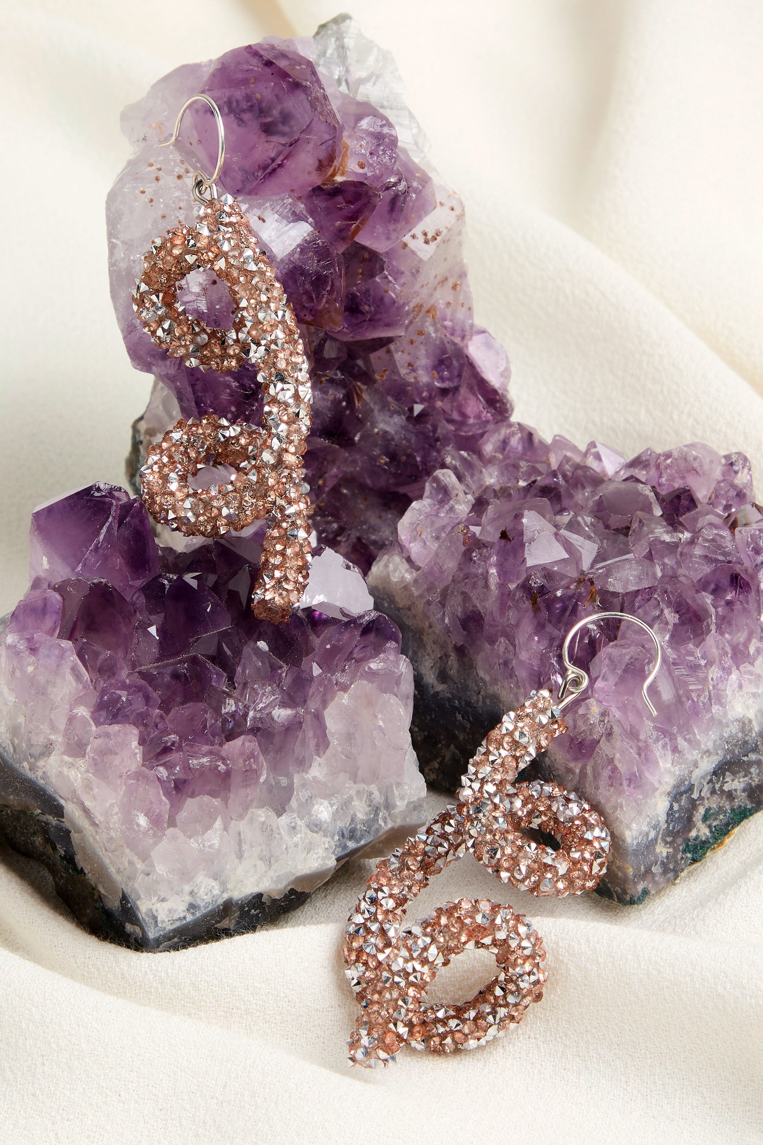 Two shimmering Disco Sketch Earrings with a twisted design elegantly sit atop clusters of amethyst crystals, set against cream fabric to create an artistic statement.