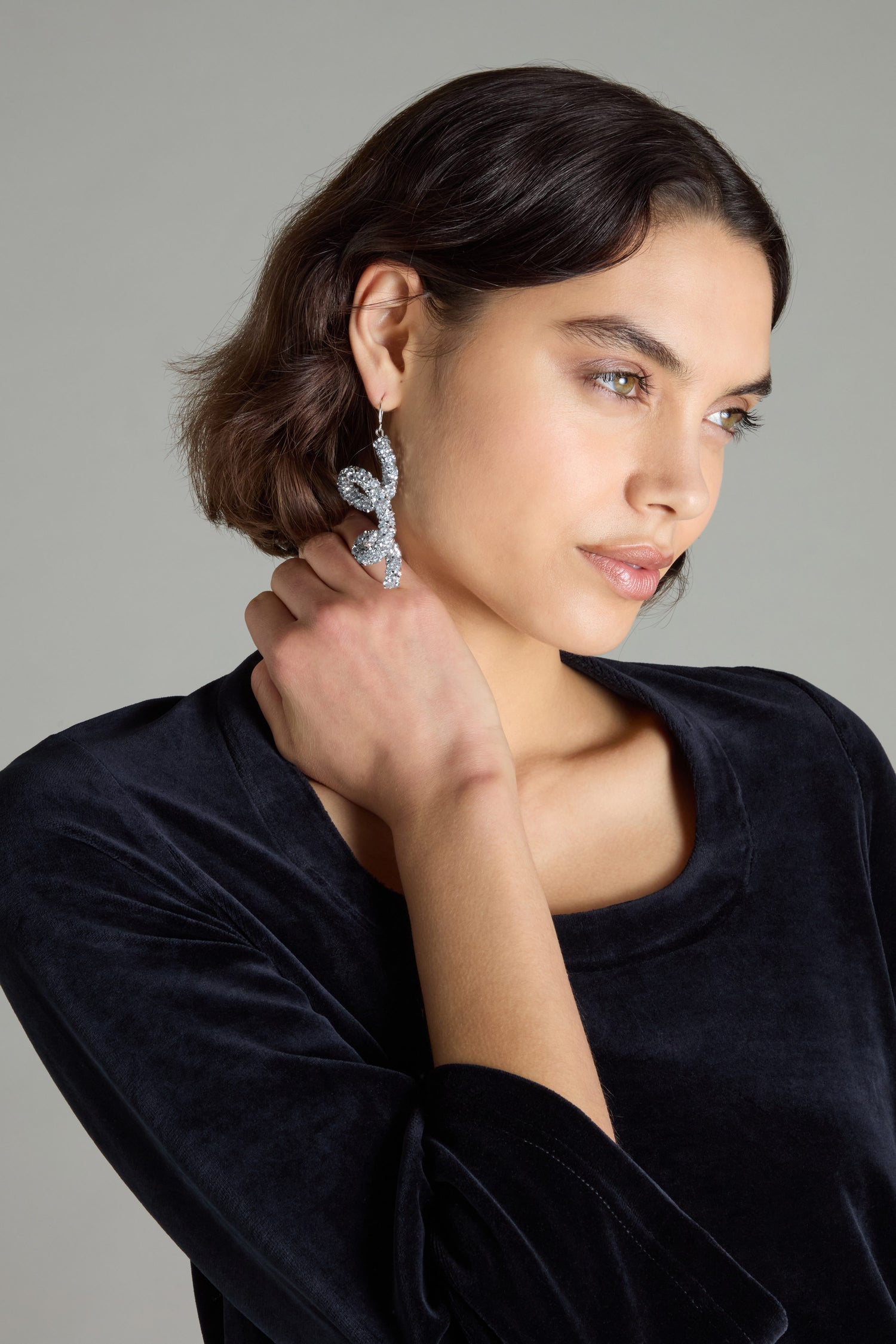 A woman with short dark hair, adorned in a black velvet top and Disco Sketch Earrings, poses gracefully with her hand on her neck against a neutral background, creating an artistic statement.