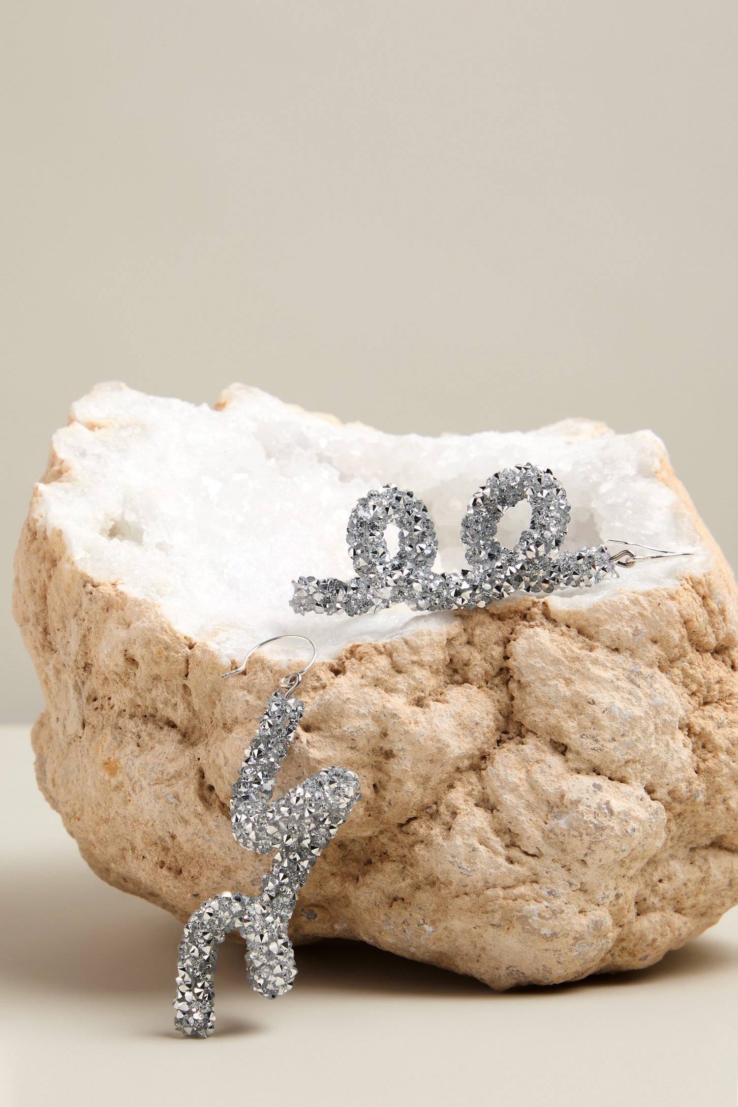 Two silver, sparkling snake-shaped earrings gleam atop a cream-colored rock, making an artistic statement reminiscent of the Disco Sketch Earrings.