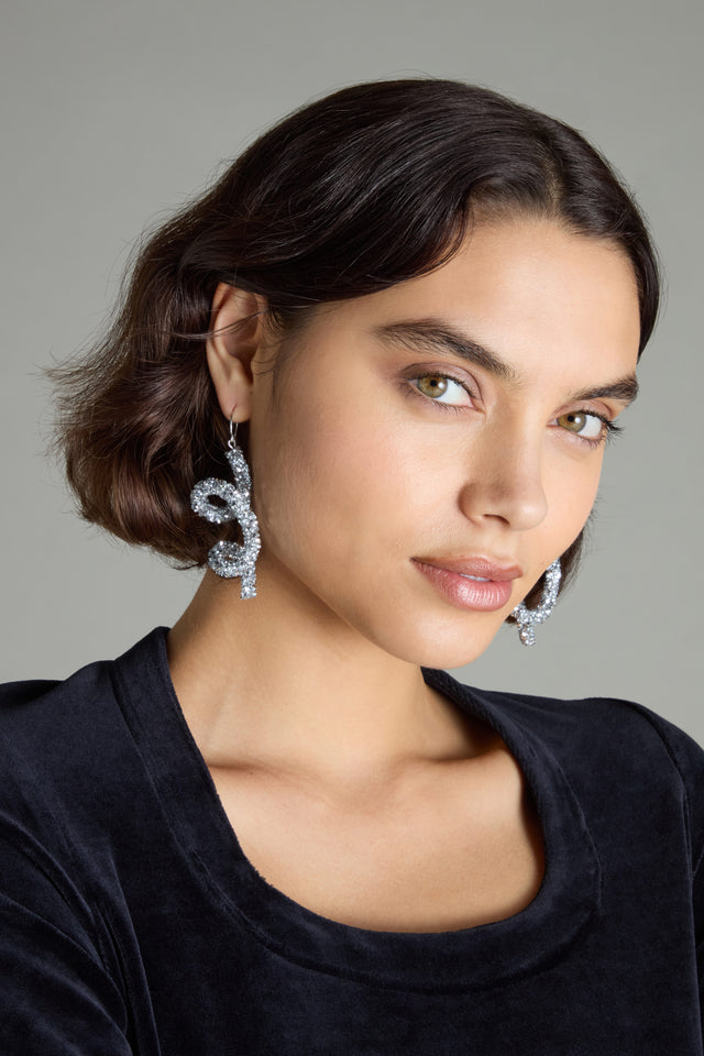 A woman with short brown hair, wearing large and ornate Disco Sketch Earrings by Samuel Coraux and a black top, makes an artistic statement as she gazes into the camera against a plain backdrop.