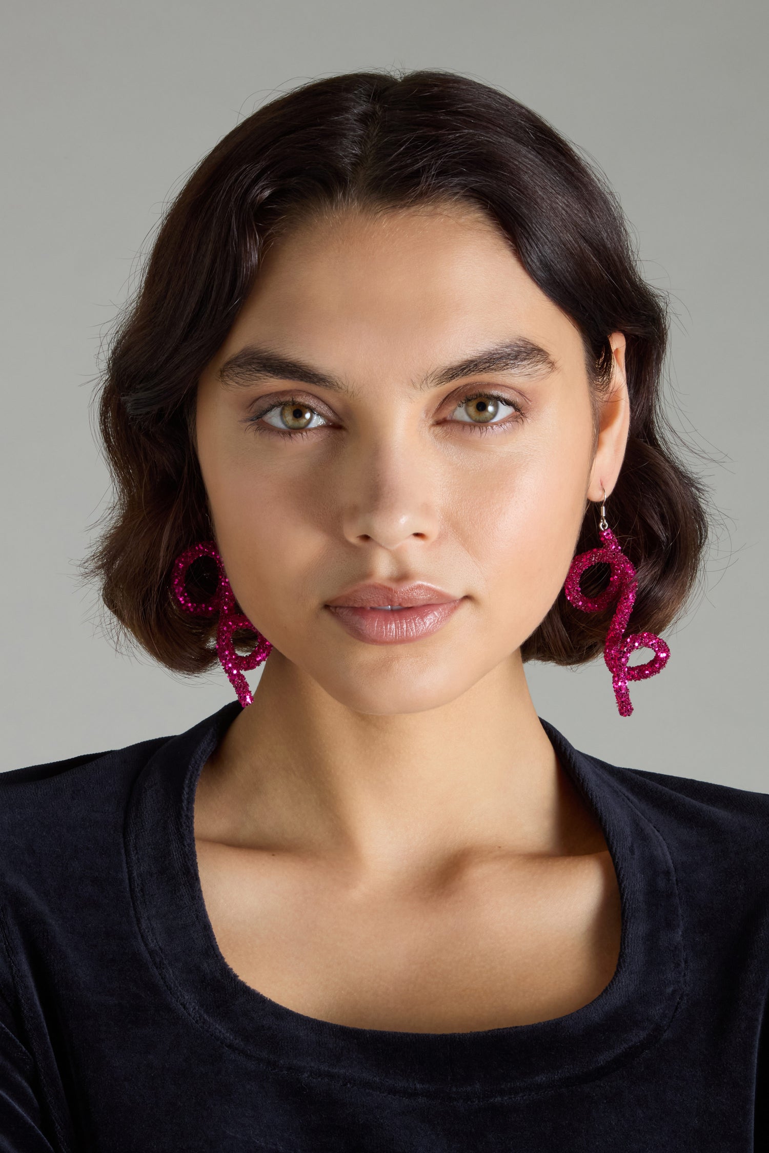 A person with short dark hair, exuding avant-garde glamour in their pink, spiral-shaped Pink Disco Sketch Earrings and a black top.