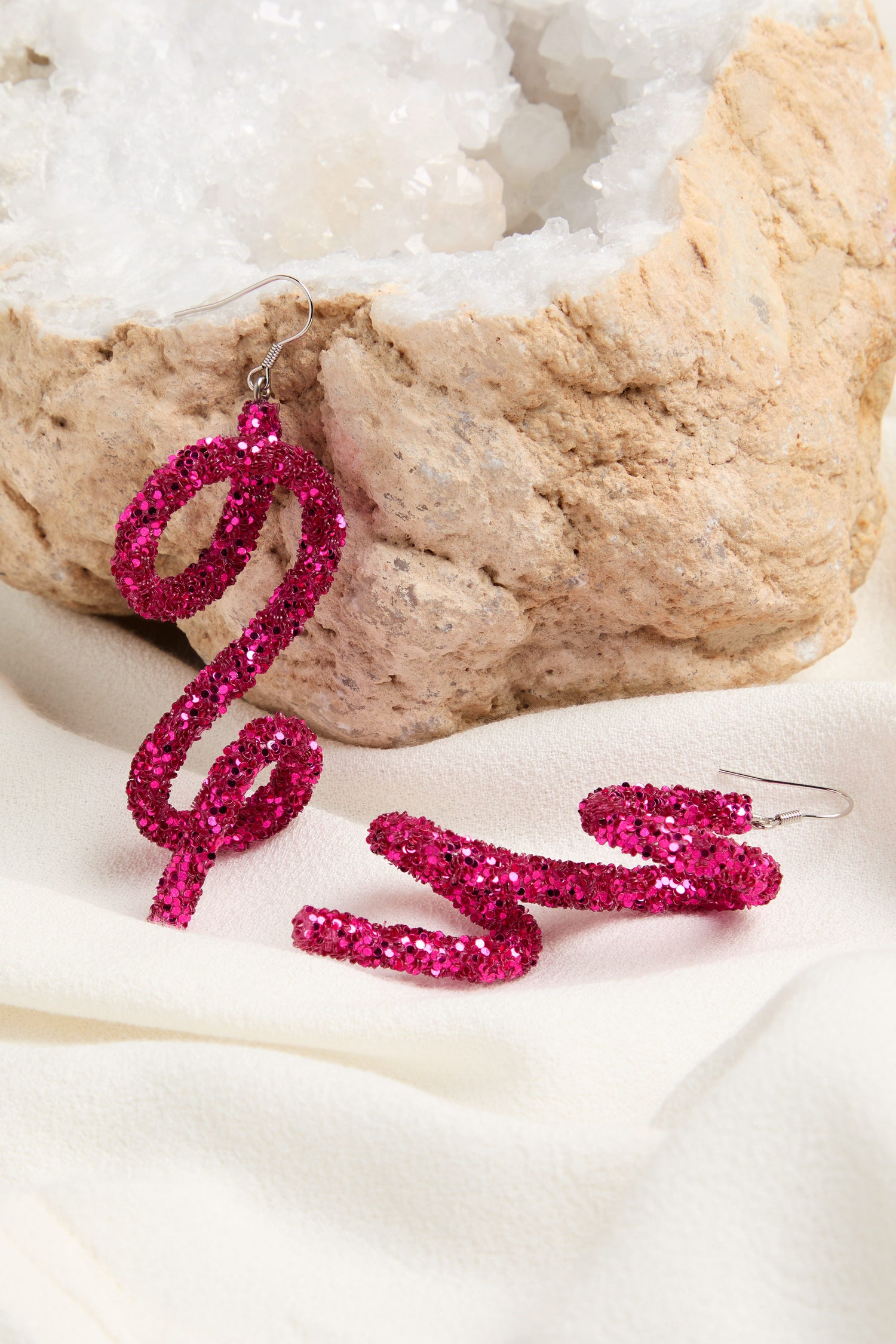 The Pink Disco Sketch Earrings, embodying avant-garde glamour, are displayed against a rough rock and white fabric background.
