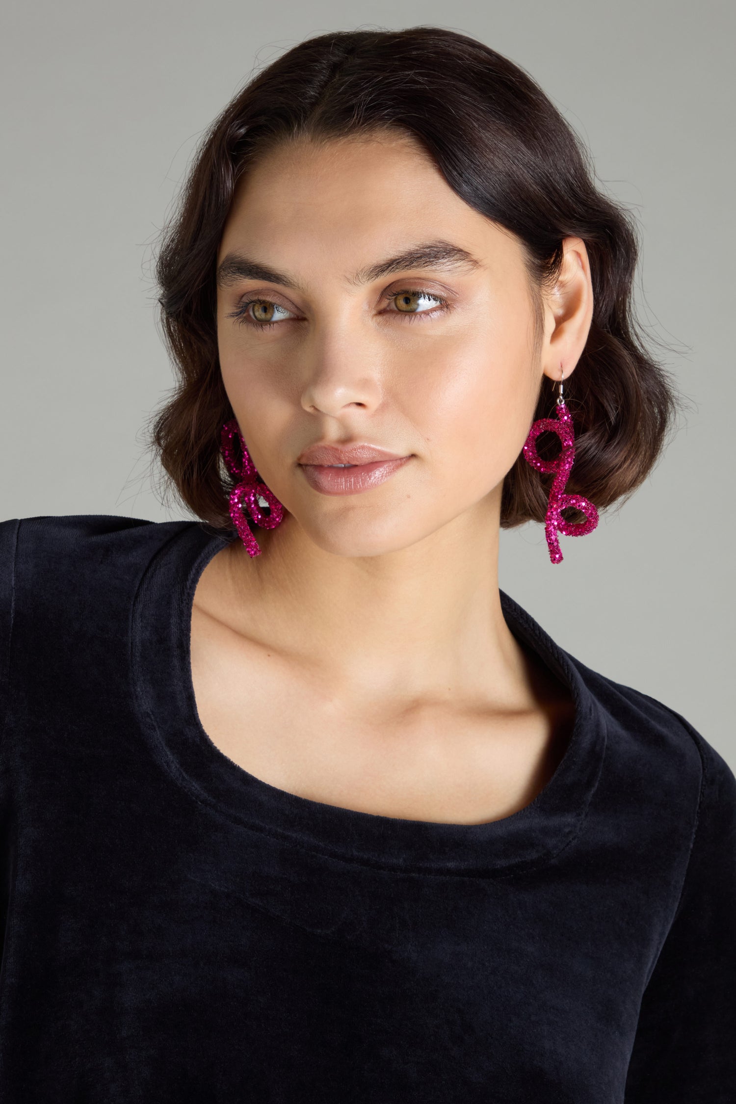 A person with short dark hair, exuding avant-garde glamour in their black top, wears Pink Disco Sketch Earrings by Samuel Coraux, looking slightly to the side.