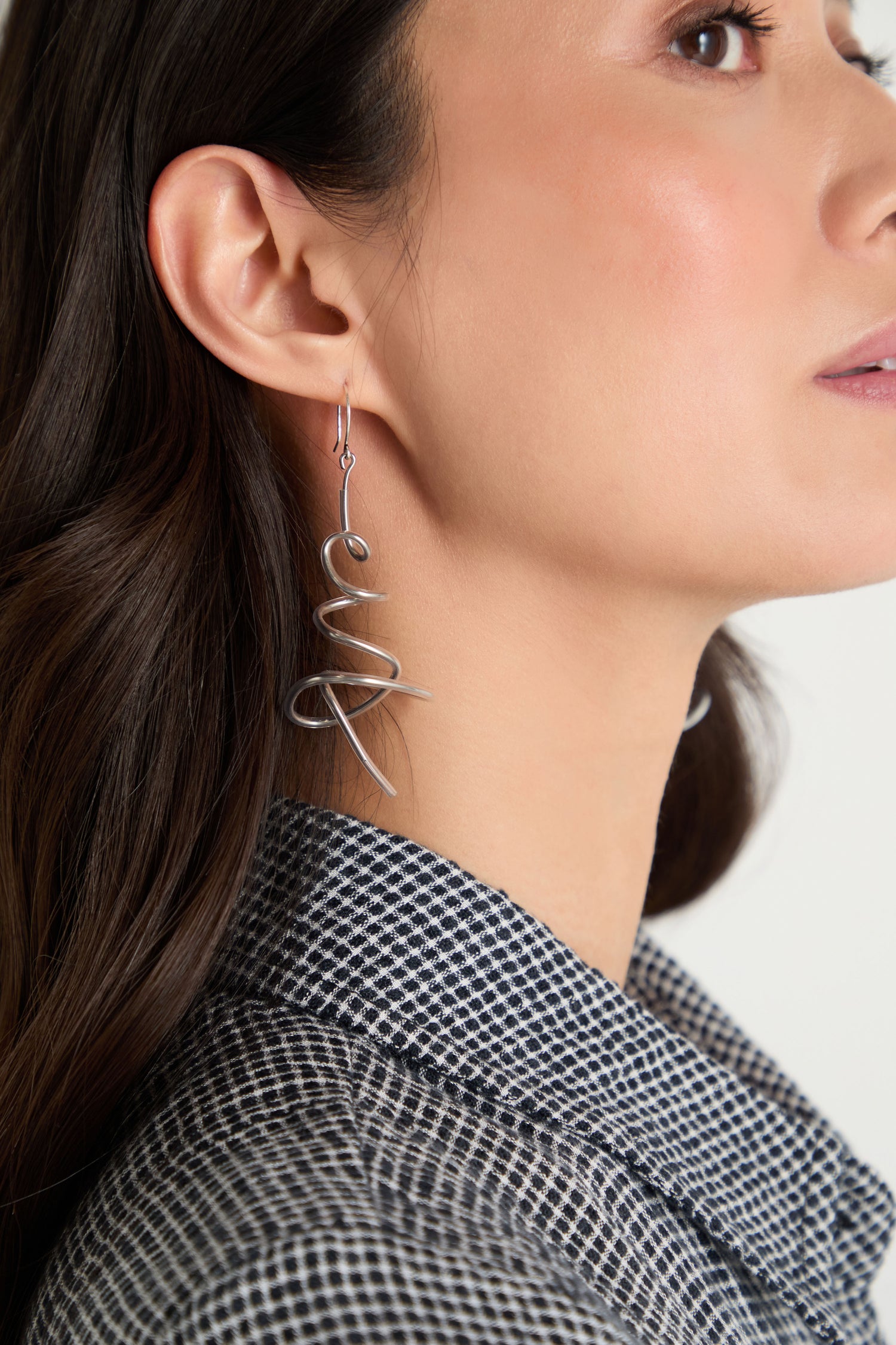 Close-up of a person wearing Squiggle Earrings—silver drop earrings with a cursive shape—styled with long dark hair and a checked collared shirt for a modern jewellery look.
