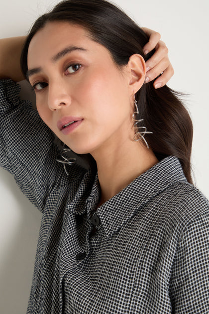 A woman with long dark hair, wearing a checked shirt and bold Squiggle Earrings, poses with one hand behind her head and looks at the camera.