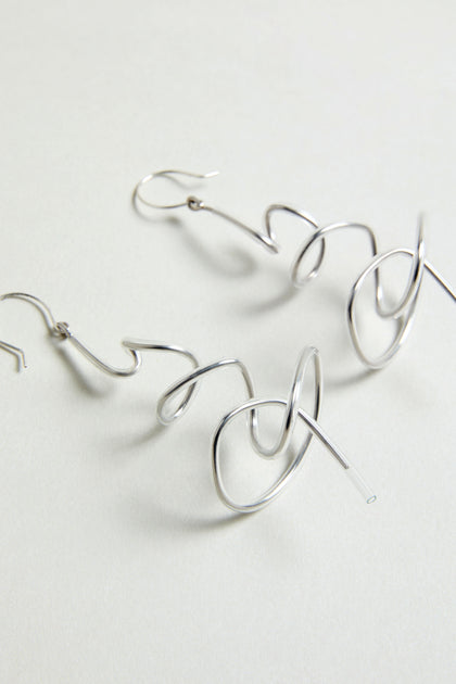 The Squiggle Earrings are silver, abstract-shaped statement drops displayed on a light neutral background—perfect for contemporary style.