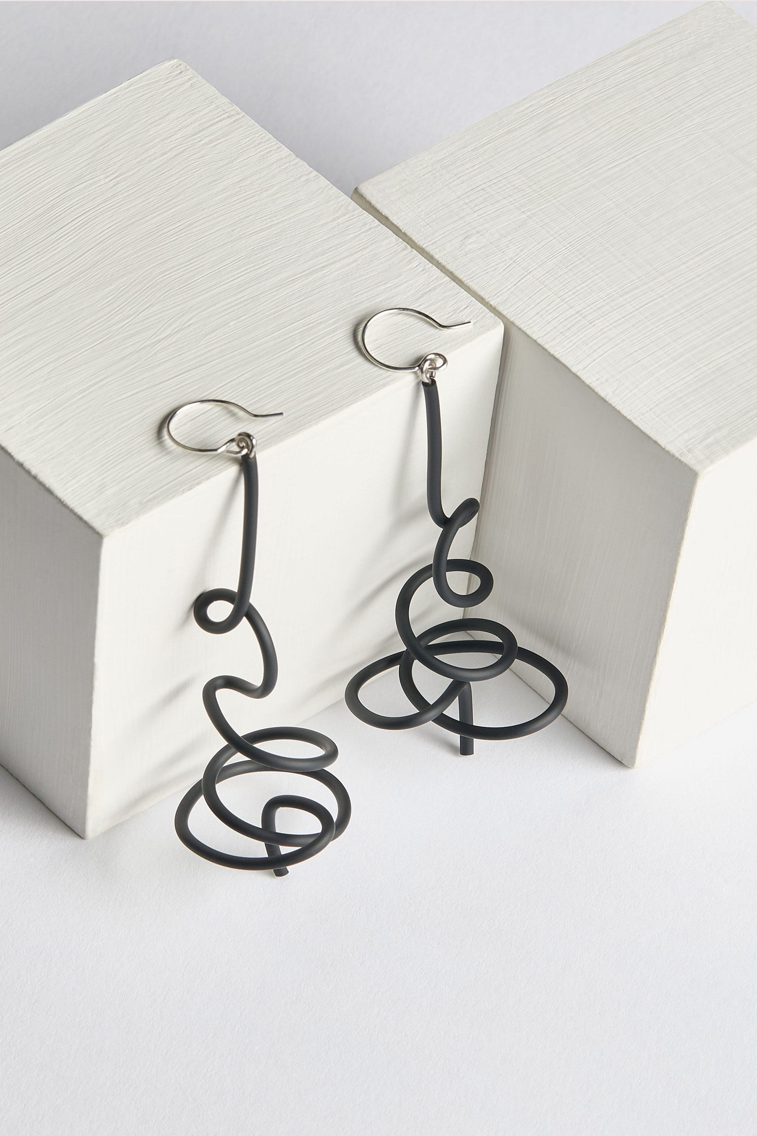 A pair of Squiggle Earrings, crafted from black metal wire and featuring Samuel Coraux's signature abstract spiral designs, are showcased on two white wooden blocks, perfectly capturing the essence of playful earrings.