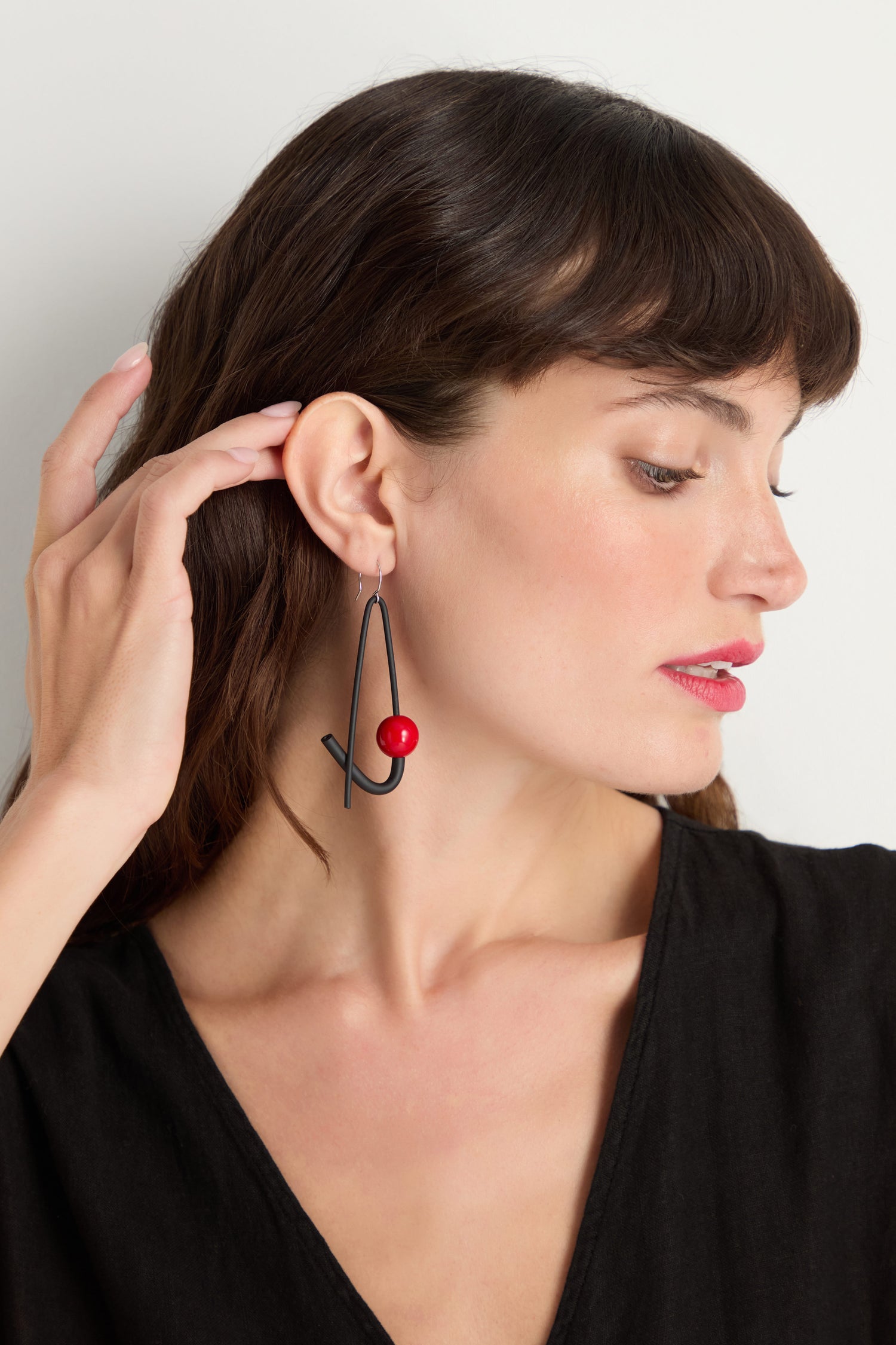 A woman in a black top wears Sphere Earrings—large geometric earrings with a red bead. Handmade in Paris, these sculptural earrings add an artistic touch to her elegant style as she looks to the side, hand near her ear.