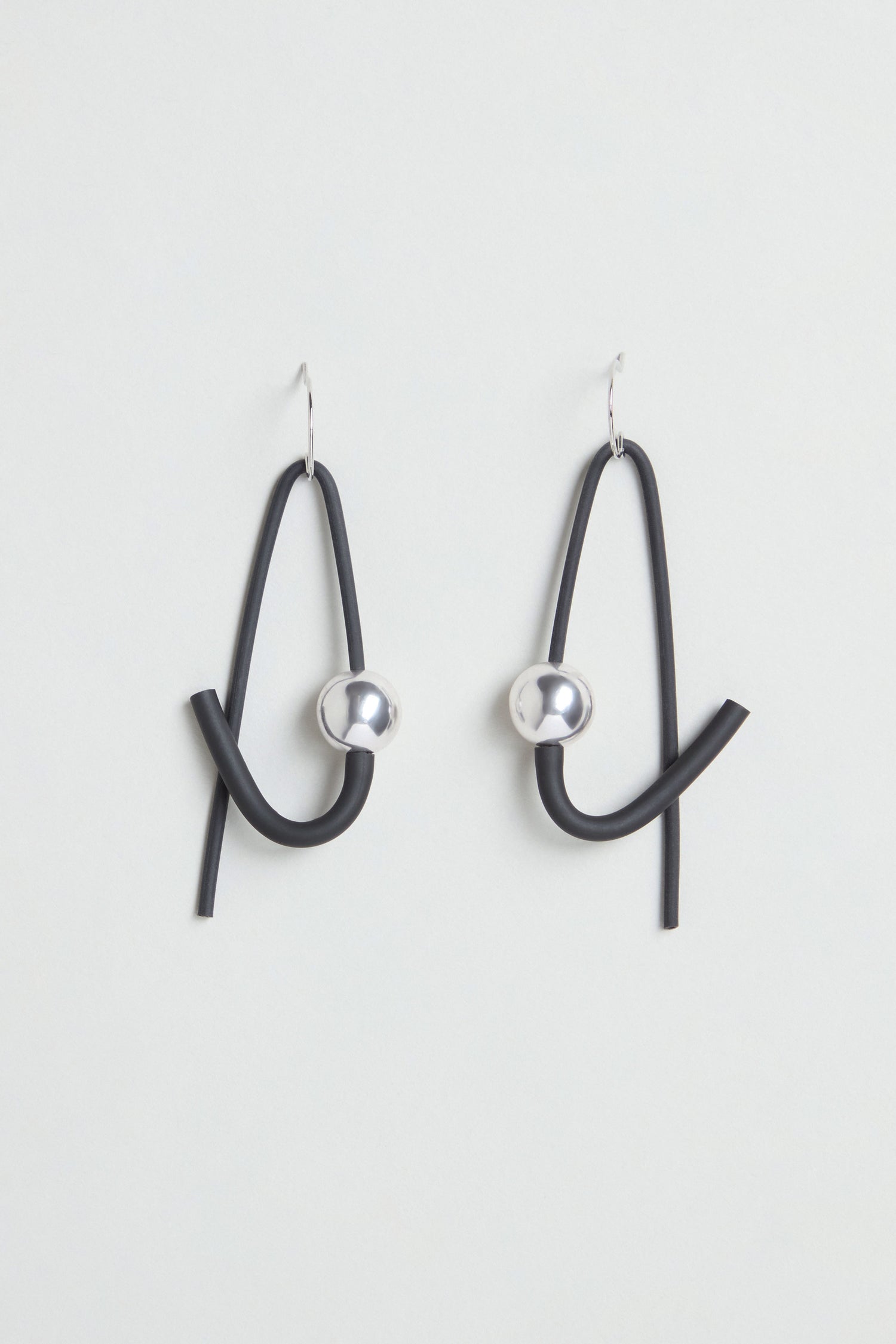 The Sphere Earrings are a pair of contemporary, handmade earrings featuring black curved loops and metallic silver spheres. Crafted in Paris, they hang from simple hooks and are displayed against a plain white background.