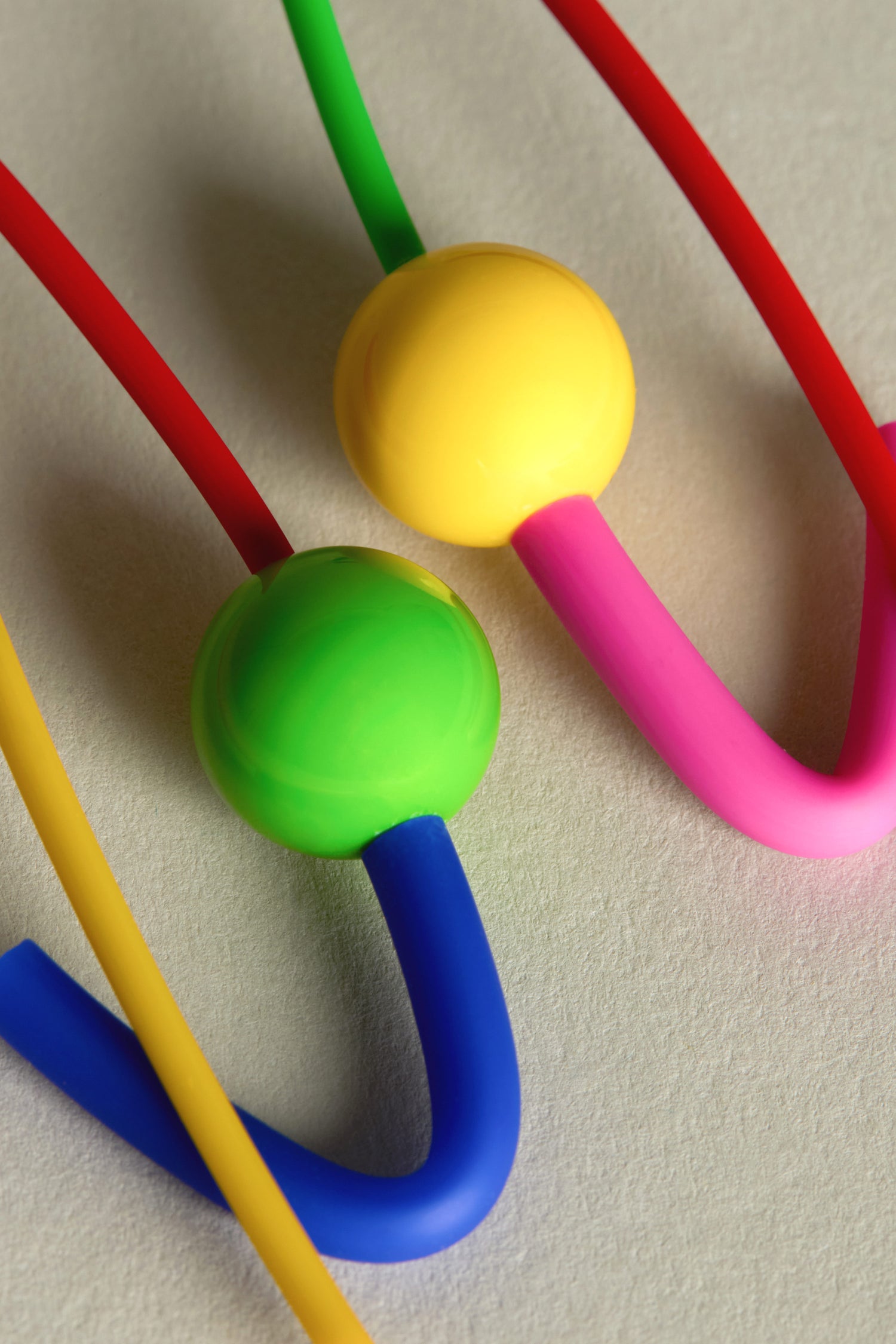 The Rainbow Sphere Earrings feature brightly colored intertwined tubes with spherical connectors, resembling Samuel Coraux jewellery. Vivid hues of red, green, yellow, pink, and blue dance across a light surface, creating a playful and artful statement piece.