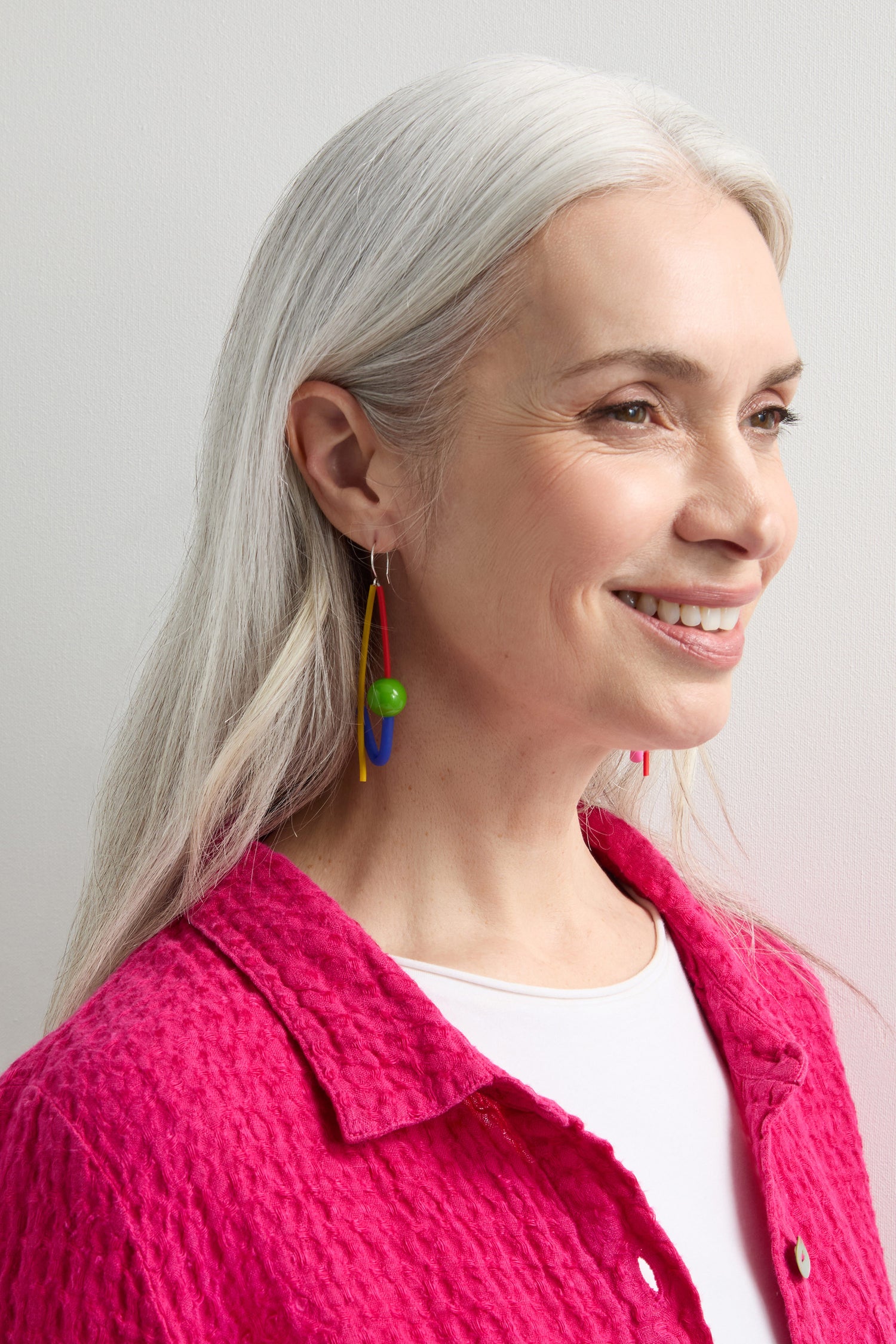 A woman with long gray hair and a bright pink textured shirt showcases the unique essence of Samuel Coraux jewellery by wearing Rainbow Sphere Earrings.