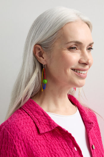 A woman with long gray hair and a bright pink textured shirt showcases the unique essence of Samuel Coraux jewellery by wearing Rainbow Sphere Earrings.