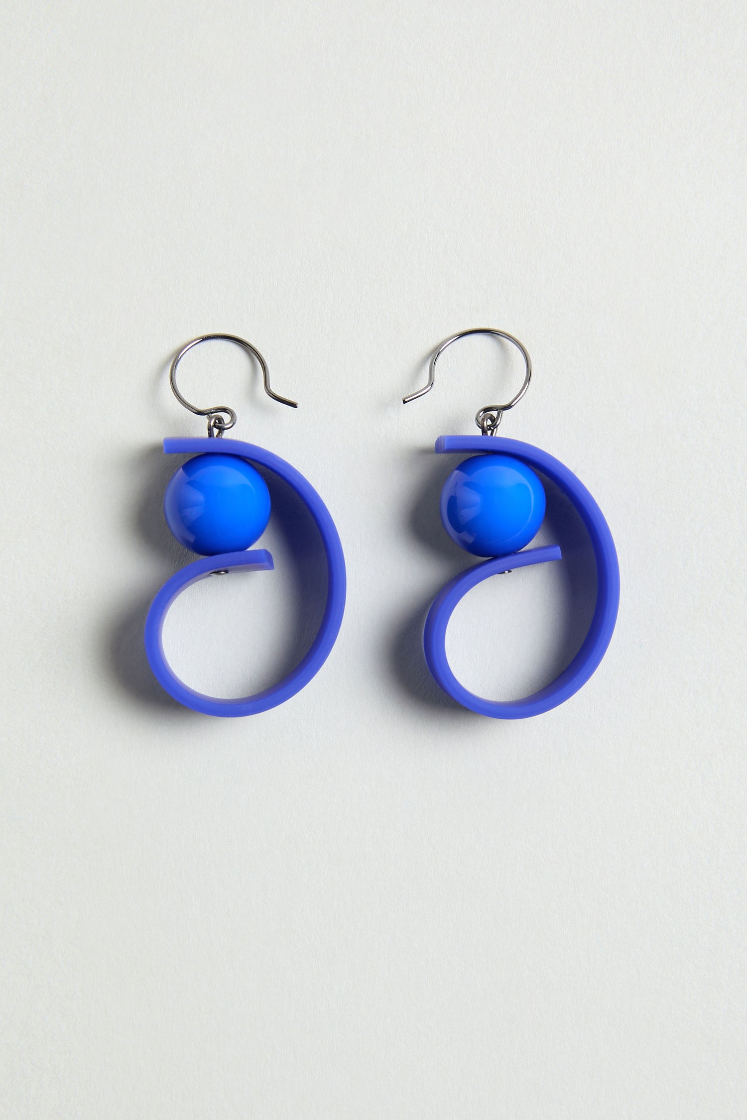 The Penseur Earrings are silver hook sculptural earrings with blue spherical beads and spiral loops, making a bold statement piece, shown on a light background.