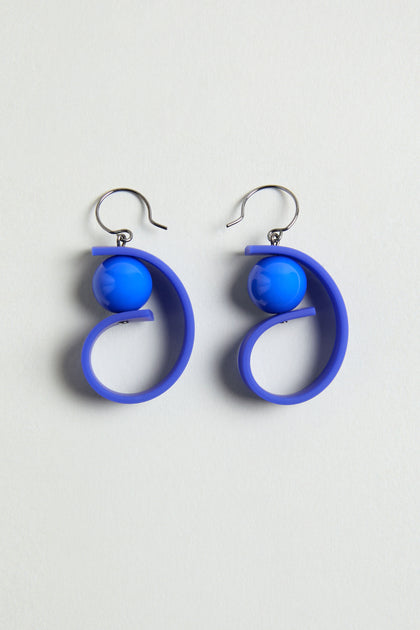The Penseur Earrings are silver hook sculptural earrings with blue spherical beads and spiral loops, making a bold statement piece, shown on a light background.