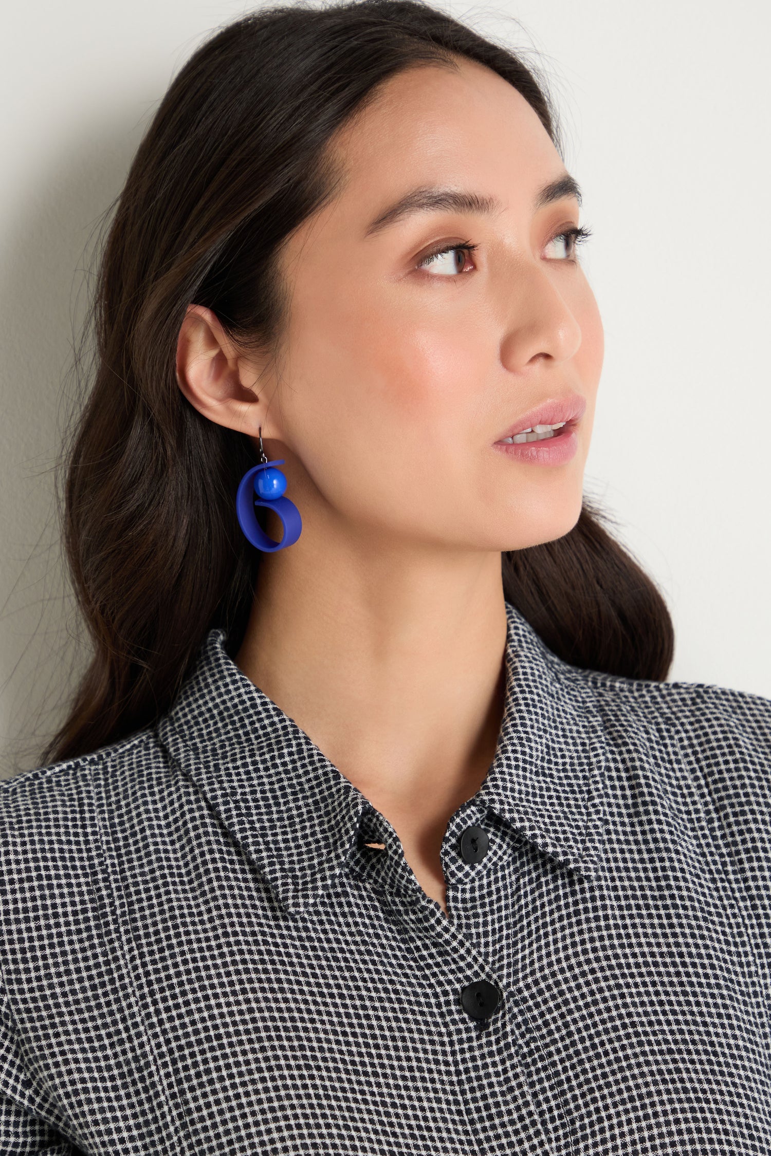 A woman with long dark hair wears a black and white checked collared shirt and Penseur Earrings in royal blue, gazing slightly to the side against a plain background.