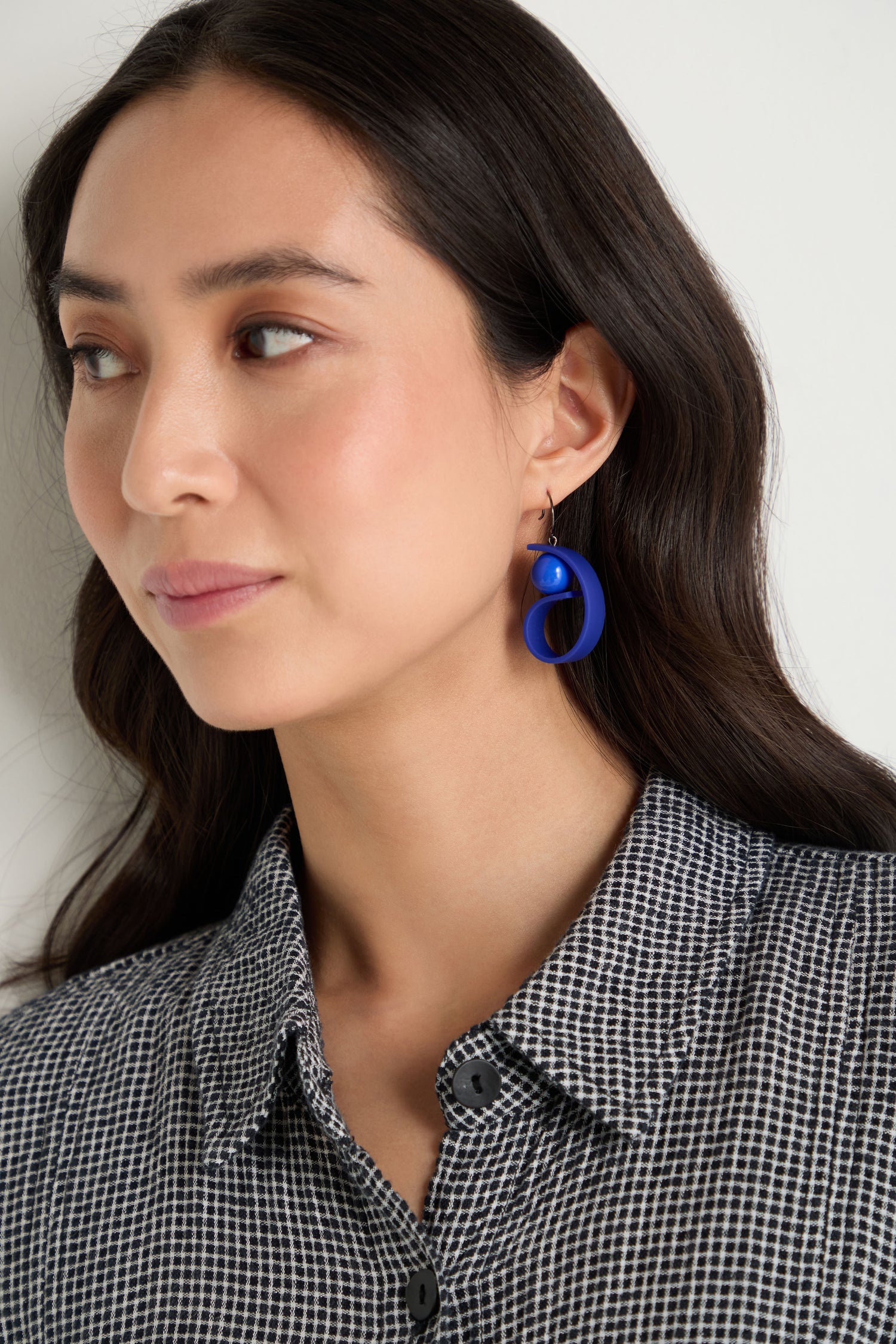 Woman with long dark hair wears the Penseur Earrings—bold, sculptural pieces in royal blue—and a black and white patterned shirt, looking slightly to the side against a plain background.