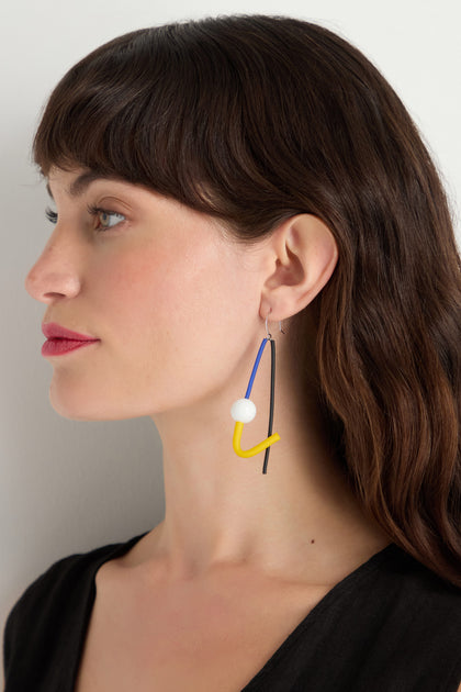 A brown-haired woman wears Kandinsky Sphere Earrings—sculptural, handmade in Paris, with blue, yellow, white, and black elements—shown in profile against a plain light background.