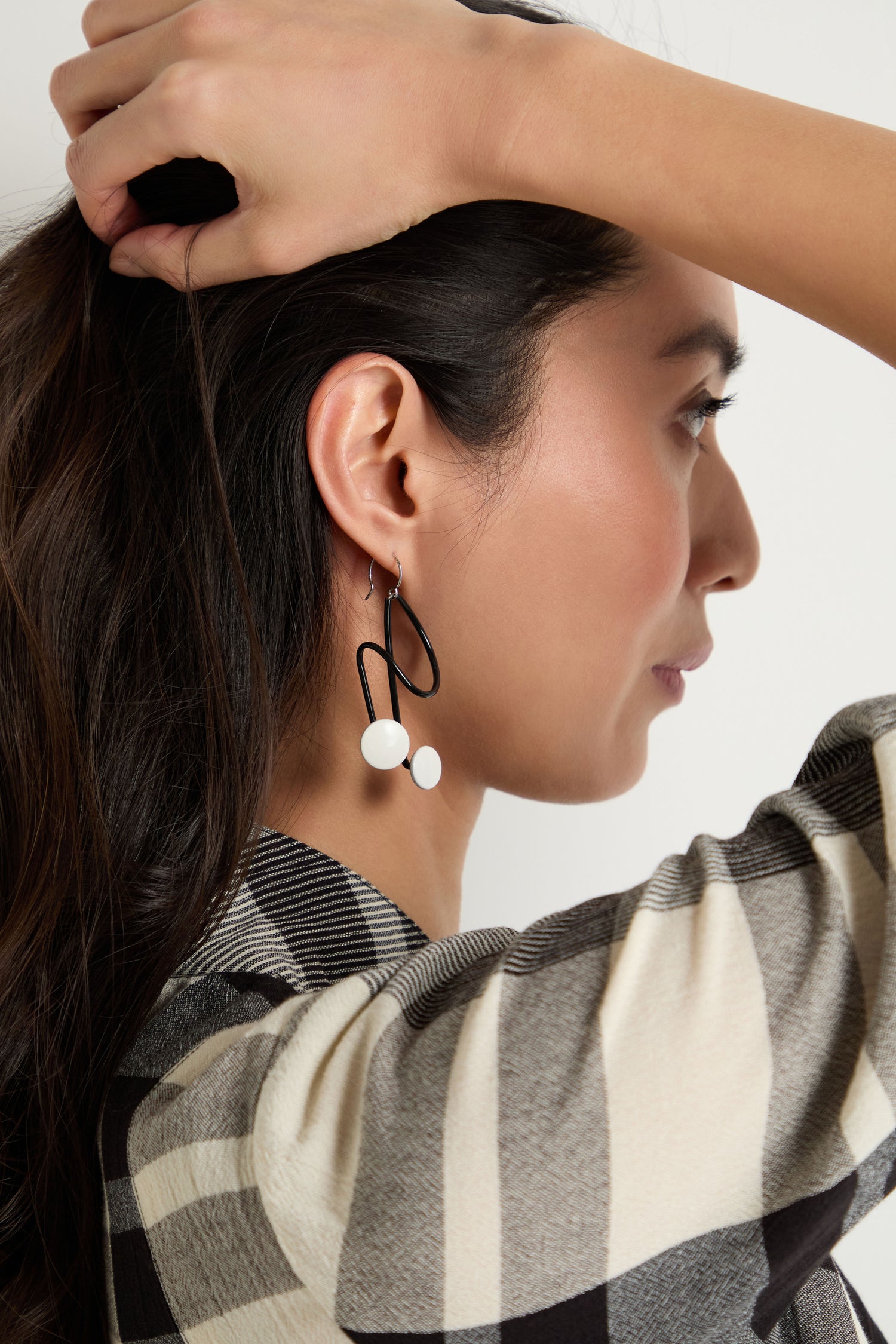 A woman in a plaid shirt holds her hair back, revealing Symphony Earrings—large geometric black and white statement pieces that exemplify contemporary jewelry.