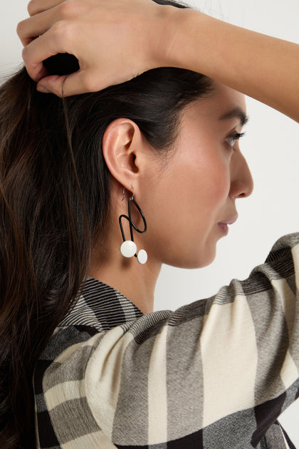 A woman in a plaid shirt holds her hair back, revealing Symphony Earrings—large geometric black and white statement pieces that exemplify contemporary jewelry.