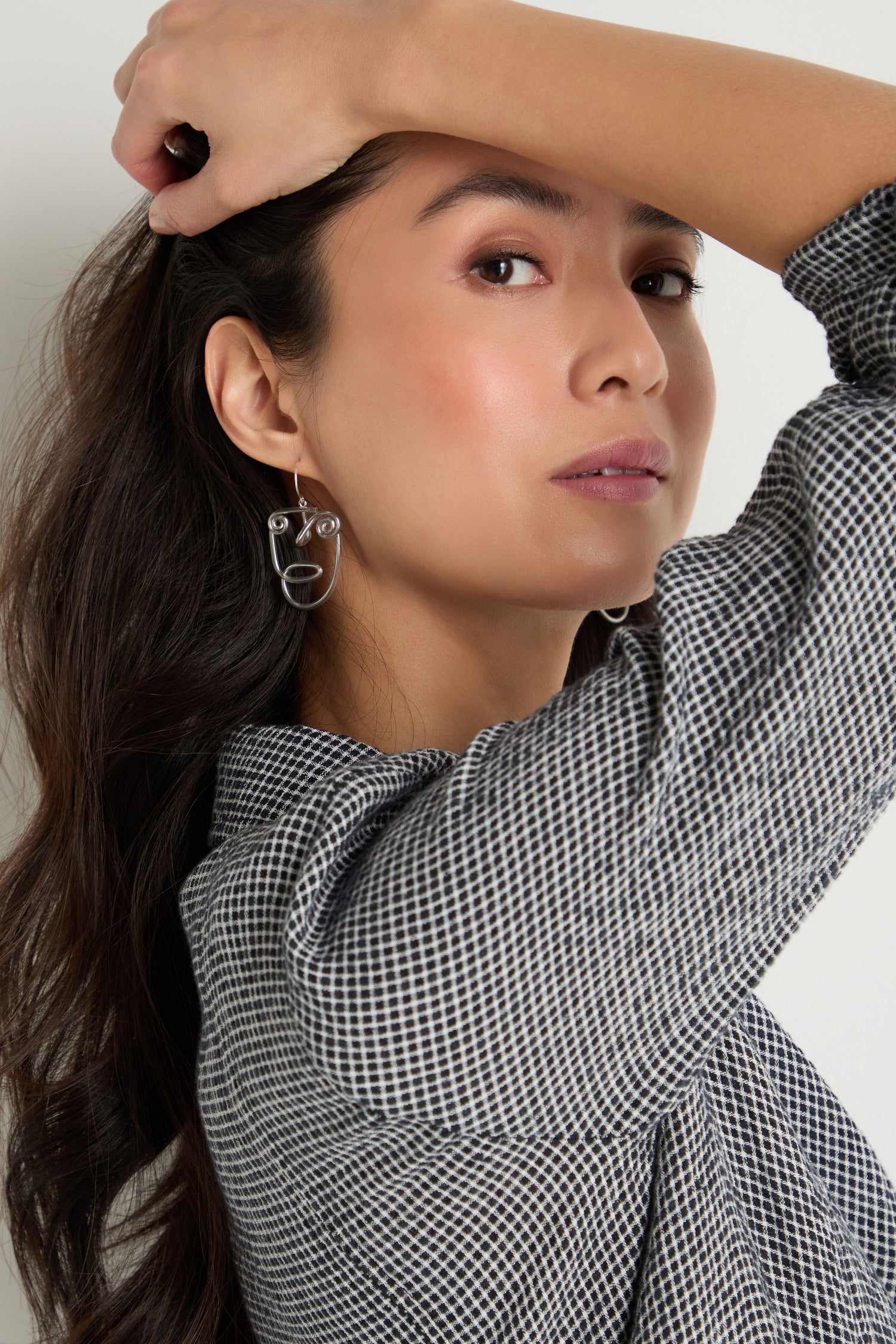 Woman with long dark hair wears a black-and-white patterned shirt and sculptural jewellery, posing with her arm raised above her head. She completes her look with striking Mini Faces Earrings.