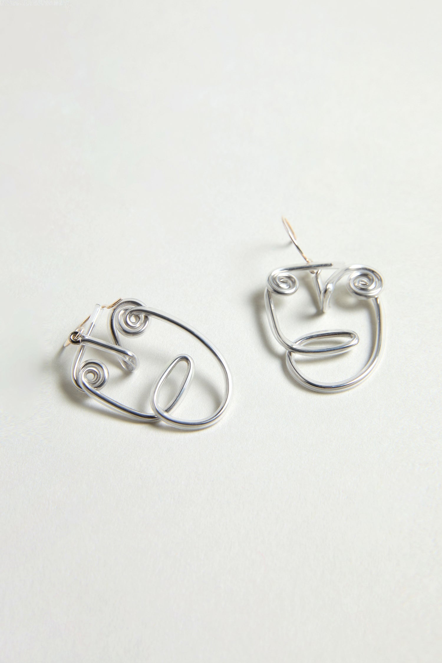 Two Mini Faces Earrings, crafted in silver with a sculptural style and shaped like abstract human faces, are displayed on a light gray surface.