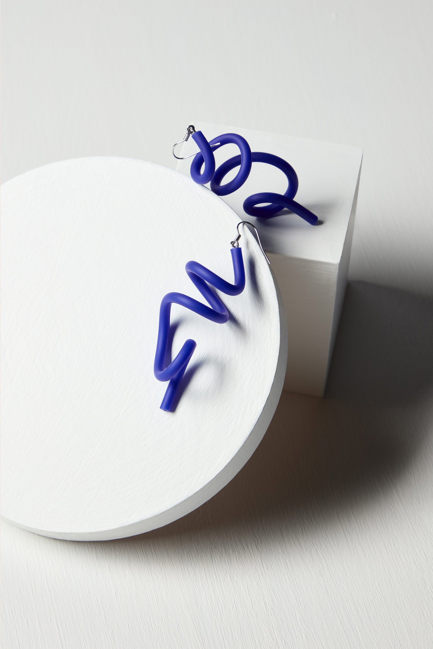 The Sketch Earrings, a pair of blue, chunky sculpted beauties, are displayed on a white circular and square platform against a light background, showcasing unique contemporary jewelry design.