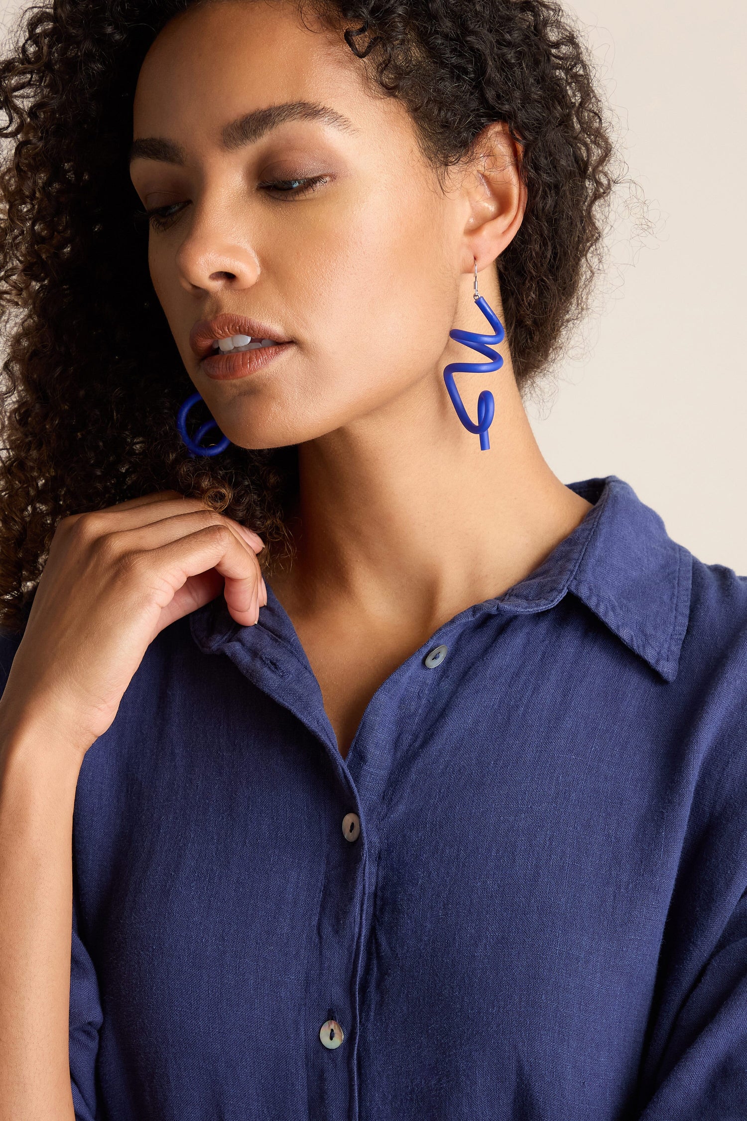 A person wearing a blue button-up shirt and contemporary jewellery, including Sketch Earrings featuring a striking blue design with a silver hook, looks downward, with one hand touching their shirt collar.