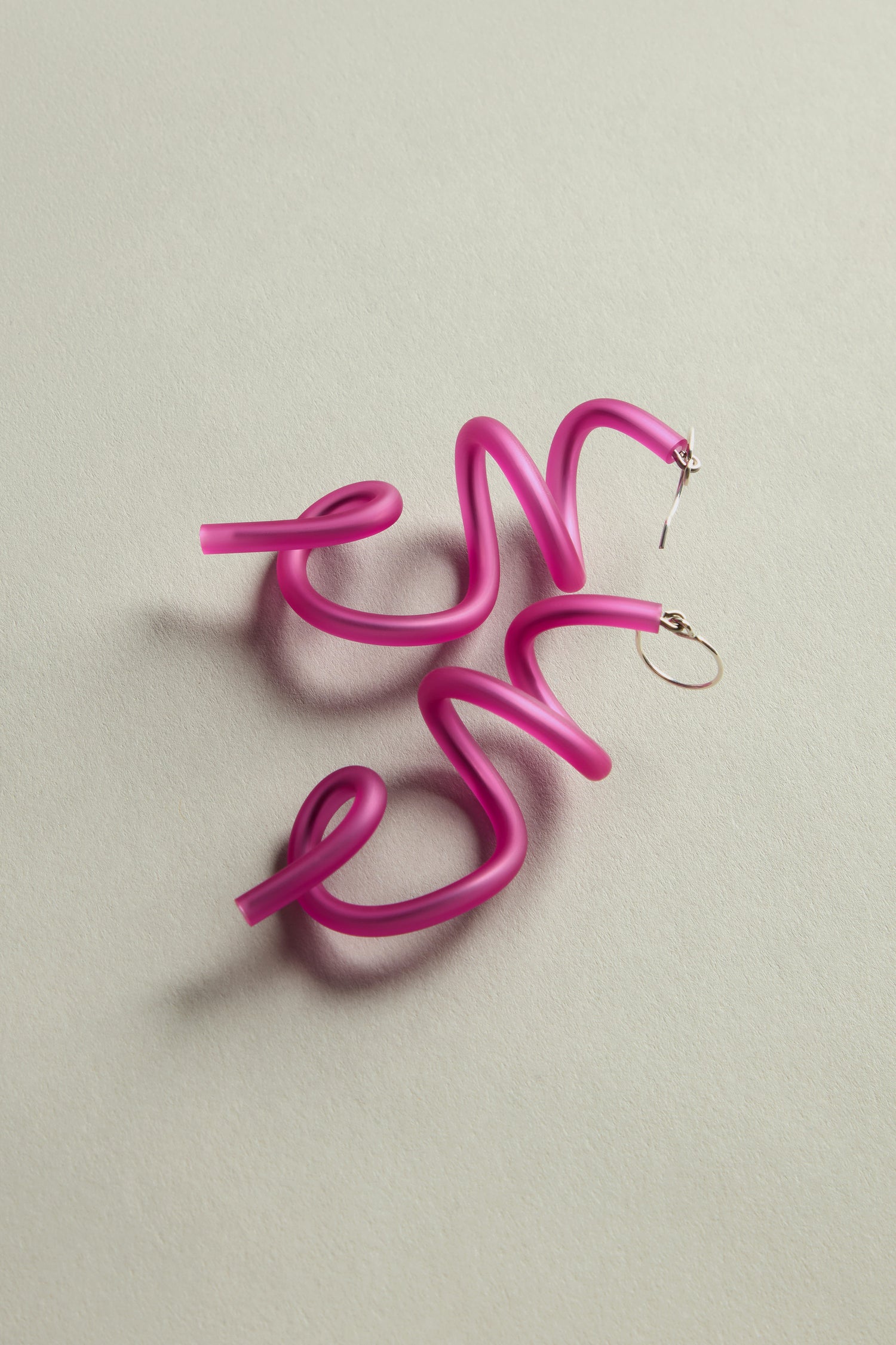 Pink, wavy, abstract hoop Sketch Earrings with gold hooks highlight contemporary design on a light gray surface.