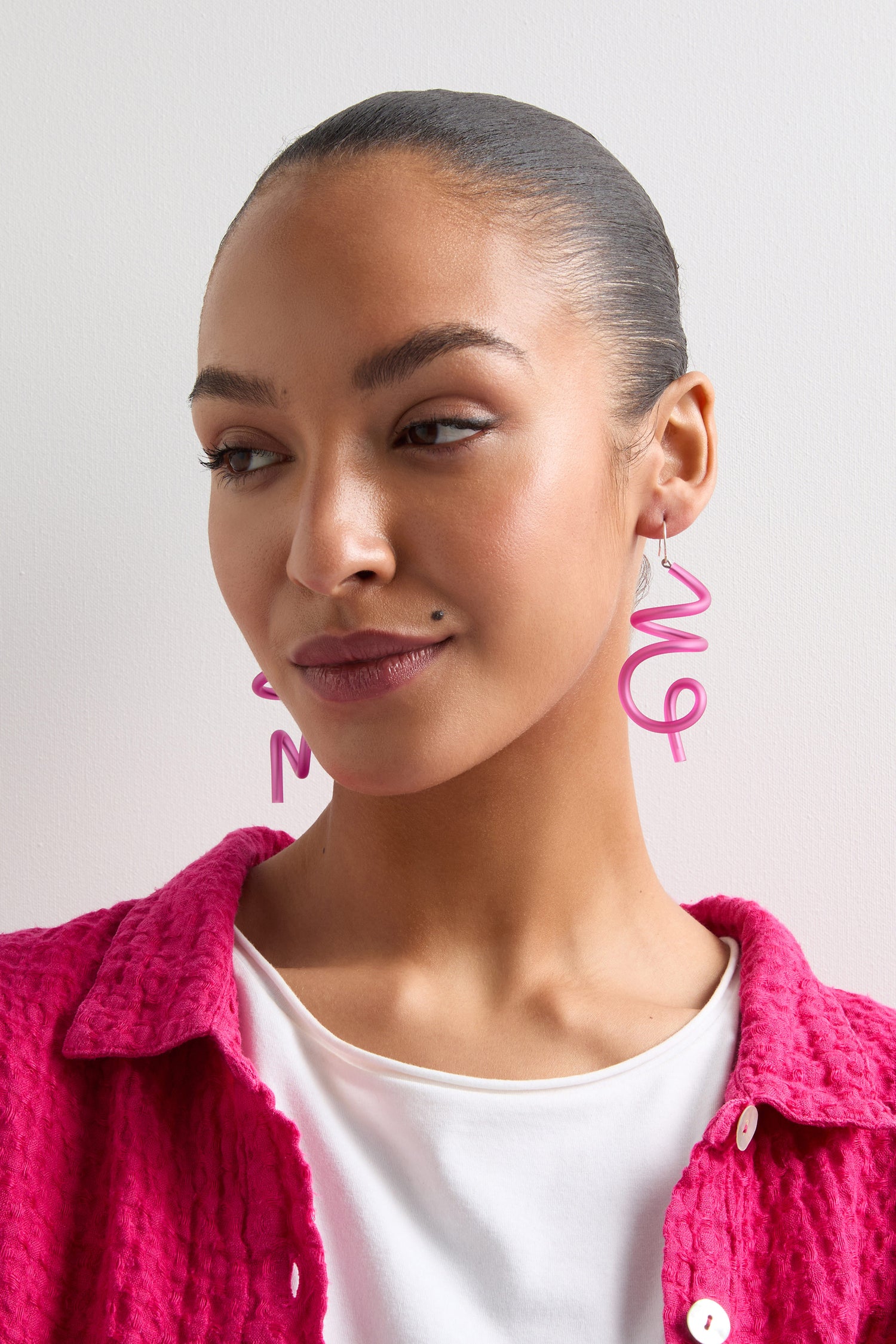 A person in a pink textured jacket and white shirt wears Sketch Earrings shaped like the word "OMG," highlighting a modern design influence.