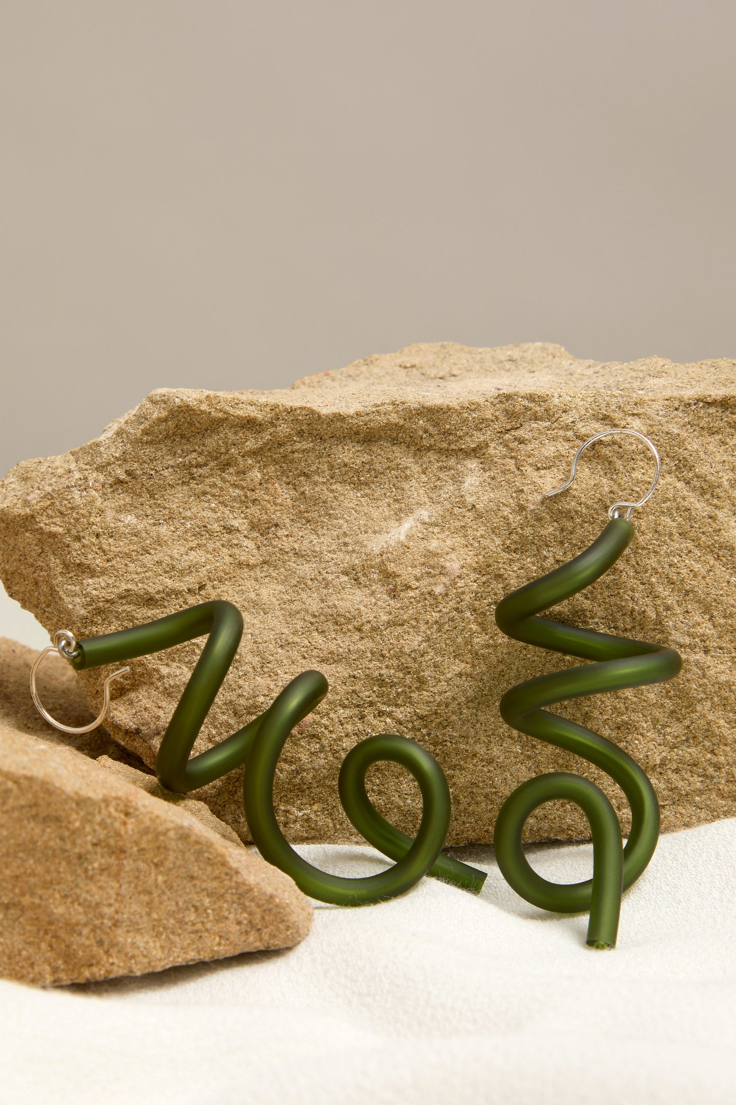 Two Sketch Earrings, in a green spiral design reminiscent of modern art, are displayed against large pieces of stone and set on a textured white fabric.