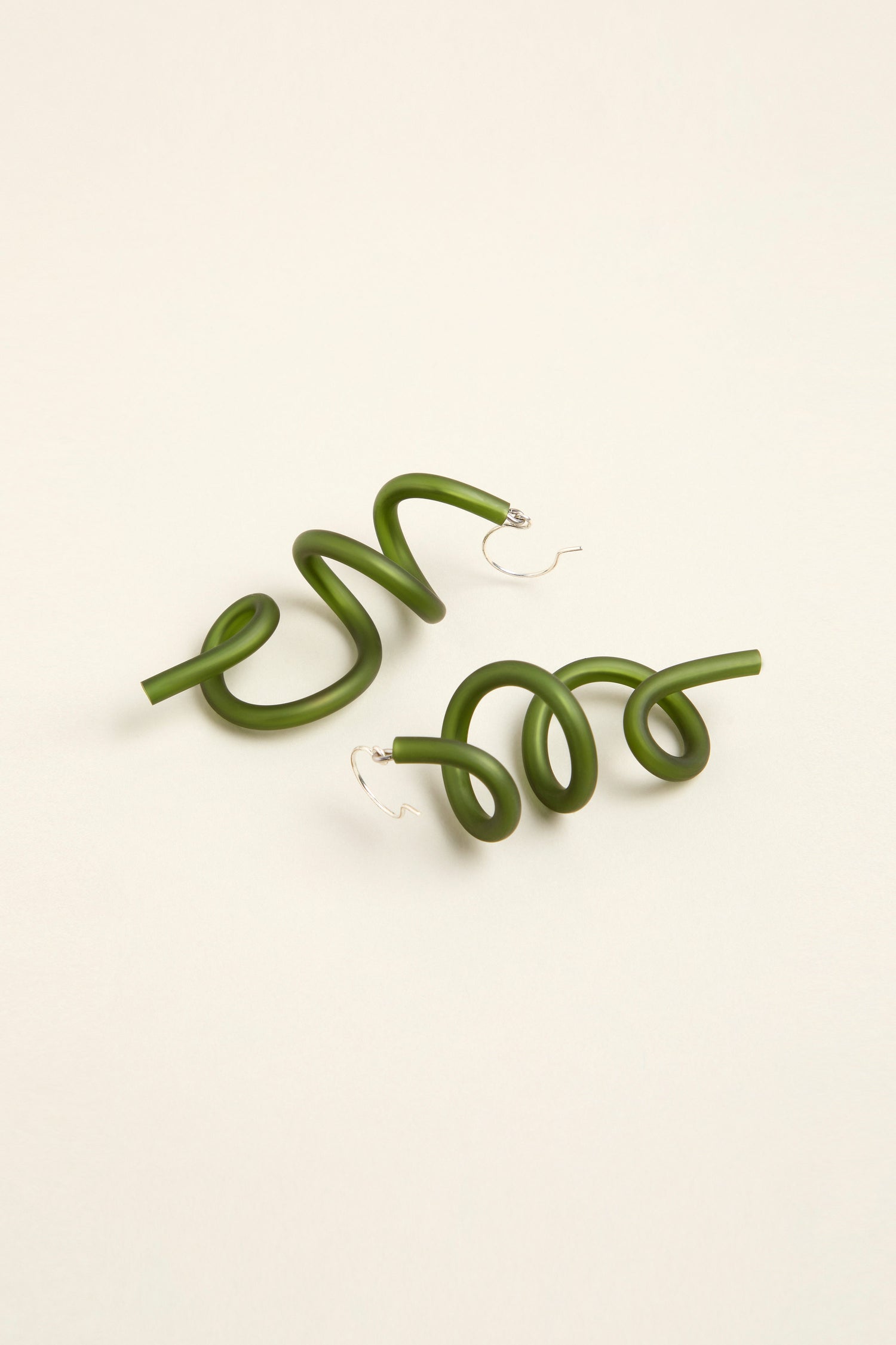 The Sketch Earrings feature green spiral hoops set against a beige background, highlighting their contemporary design.