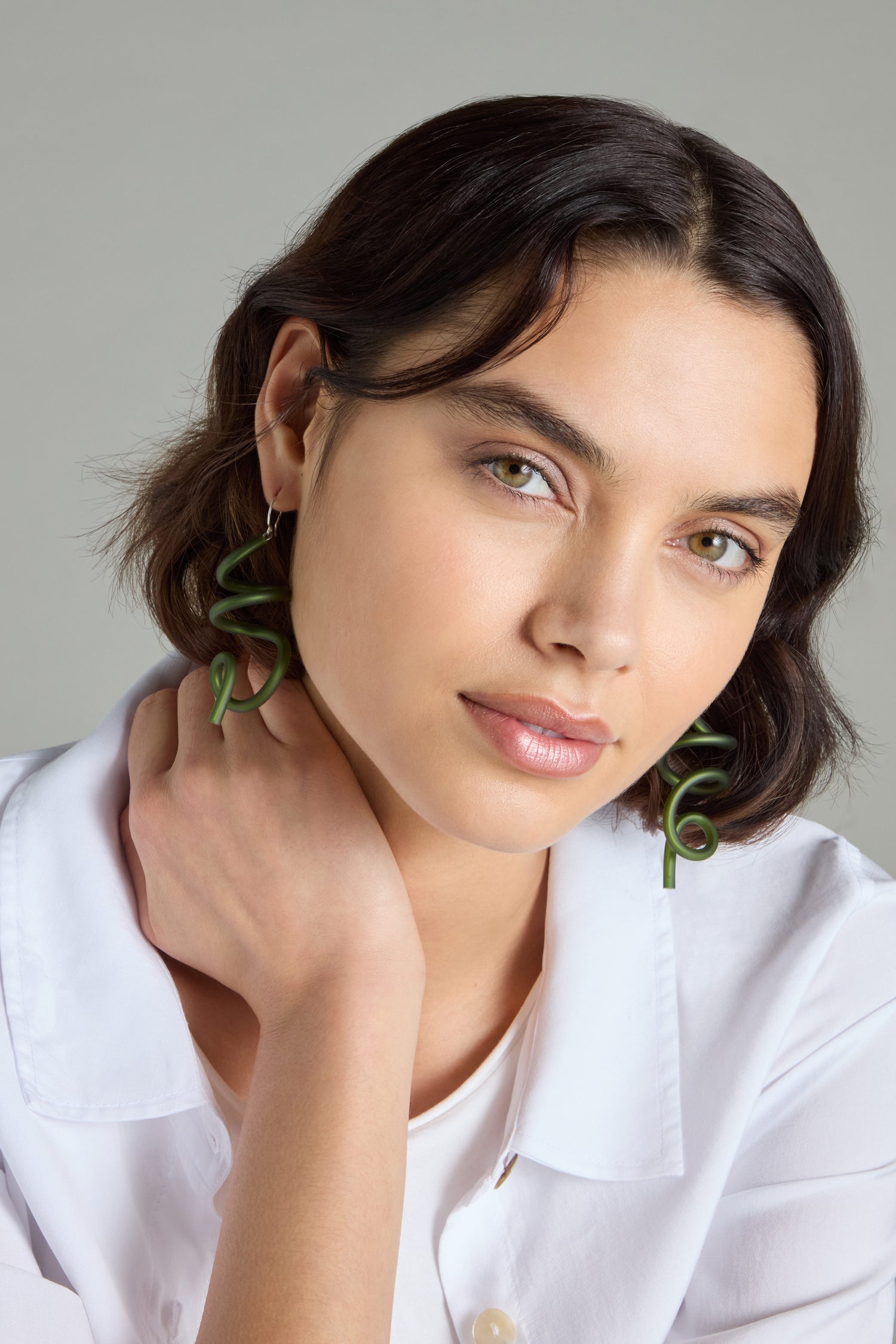 A person with short dark hair wears a white shirt and dons Sketch Earrings, which take inspiration from modern art, posing with a hand on their neck against a neutral background.