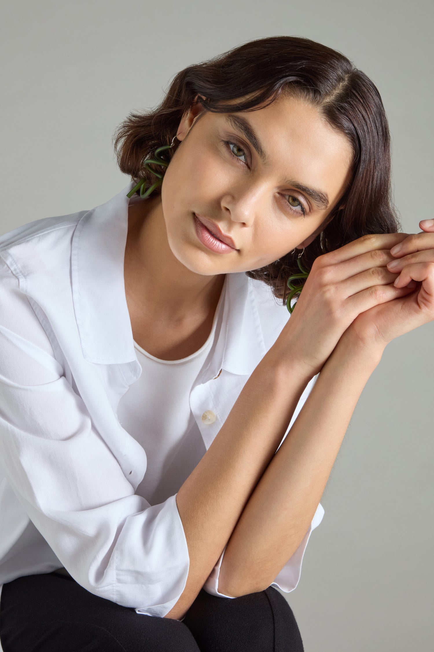 A person with short brown hair, wearing a white shirt and Sketch Earrings from the Modern Art collection, leans forward, clasping their hands.