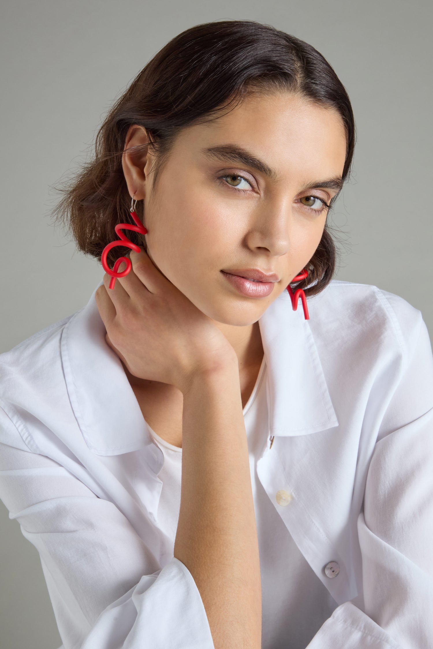 A person with short brown hair, wearing red Sketch Earrings and a white shirt, rests their chin on their hand against a background that evokes modern art's simplicity.