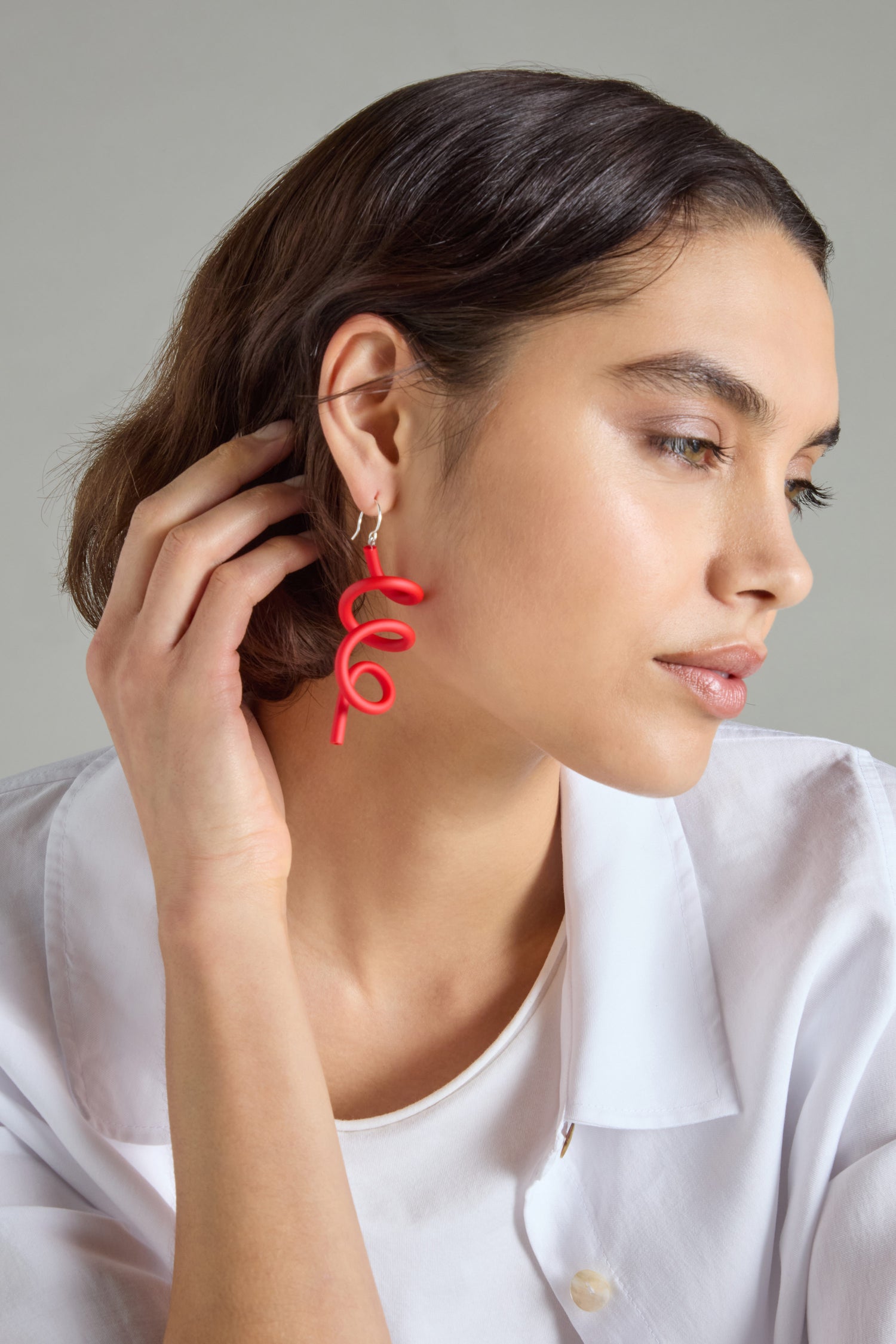 Wearing a white shirt, a person adorned in Sketch Earrings with a striking red spiral motif exudes modern elegance as they softly touch their ear, maintaining a calm expression.