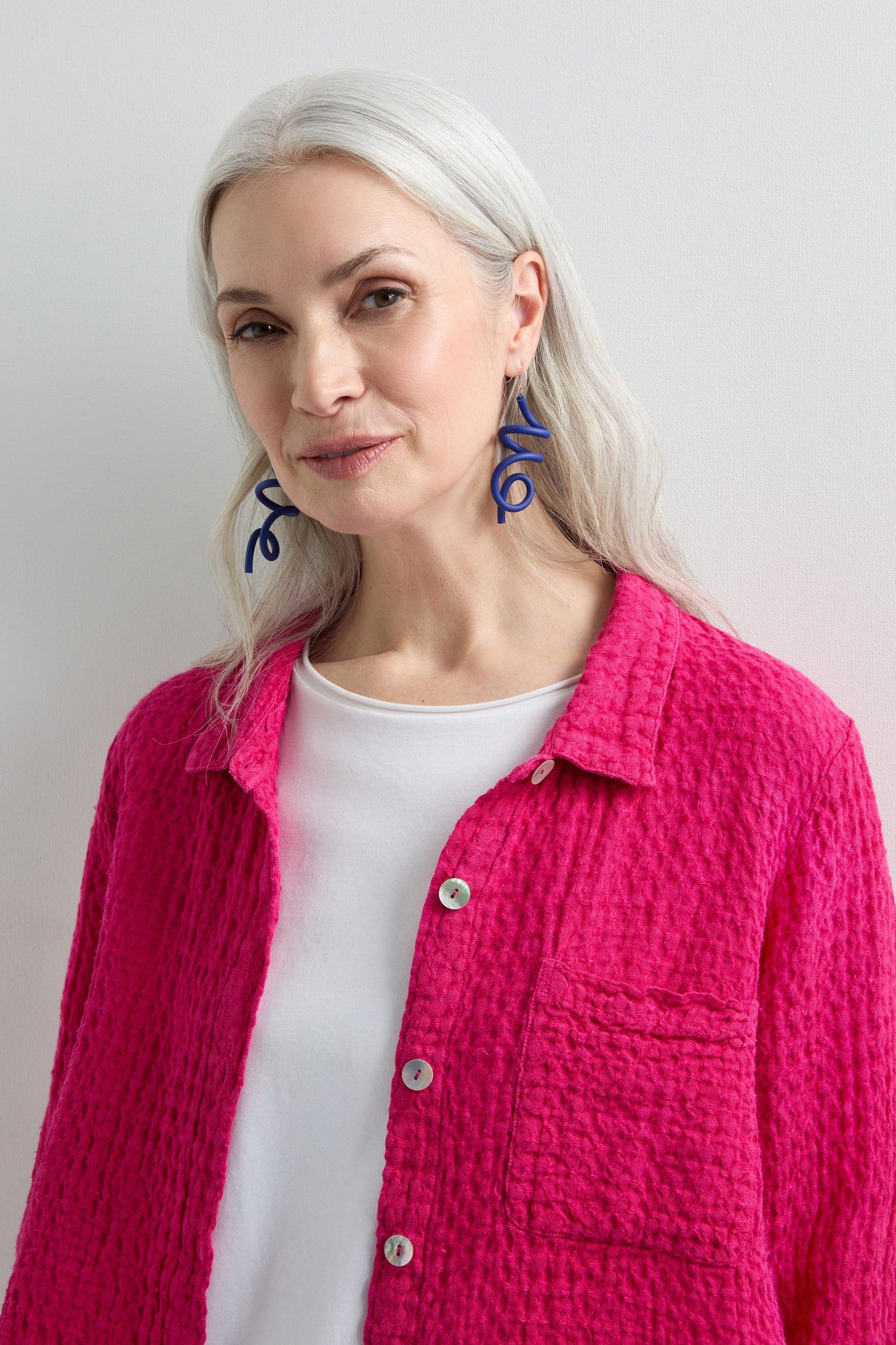 A person with long gray hair wears a bright pink textured jacket over a white top and navy Sketch Earrings, reminiscent of modern art, against a plain background.