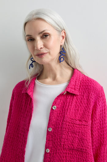 A person with long gray hair wears a bright pink textured jacket over a white top and navy Sketch Earrings, reminiscent of modern art, against a plain background.