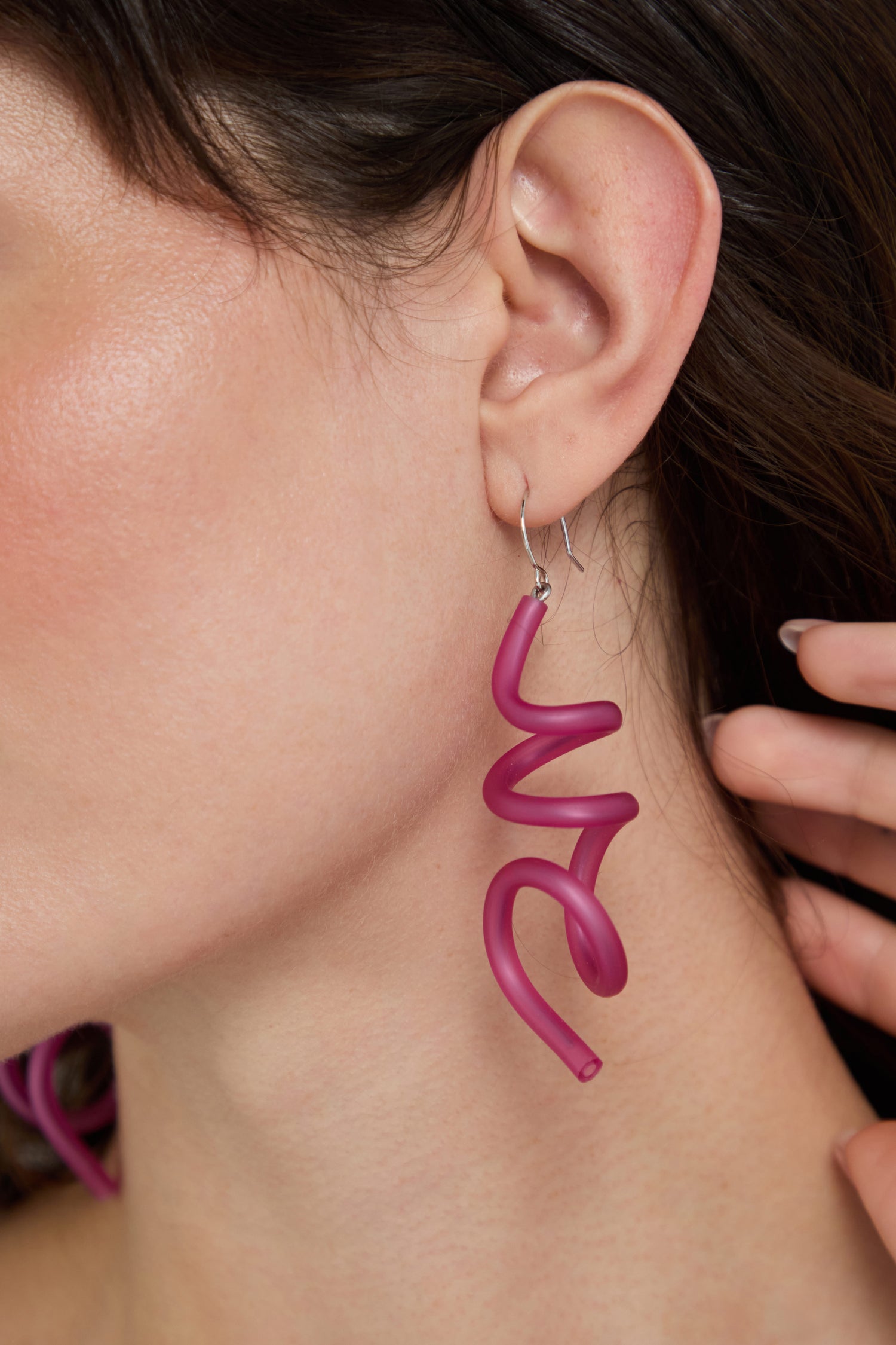 Close-up of an ear wearing large, fuchsia Sketch Earrings in an abstract sculptural design, with a hand softly touching hair near the ear.