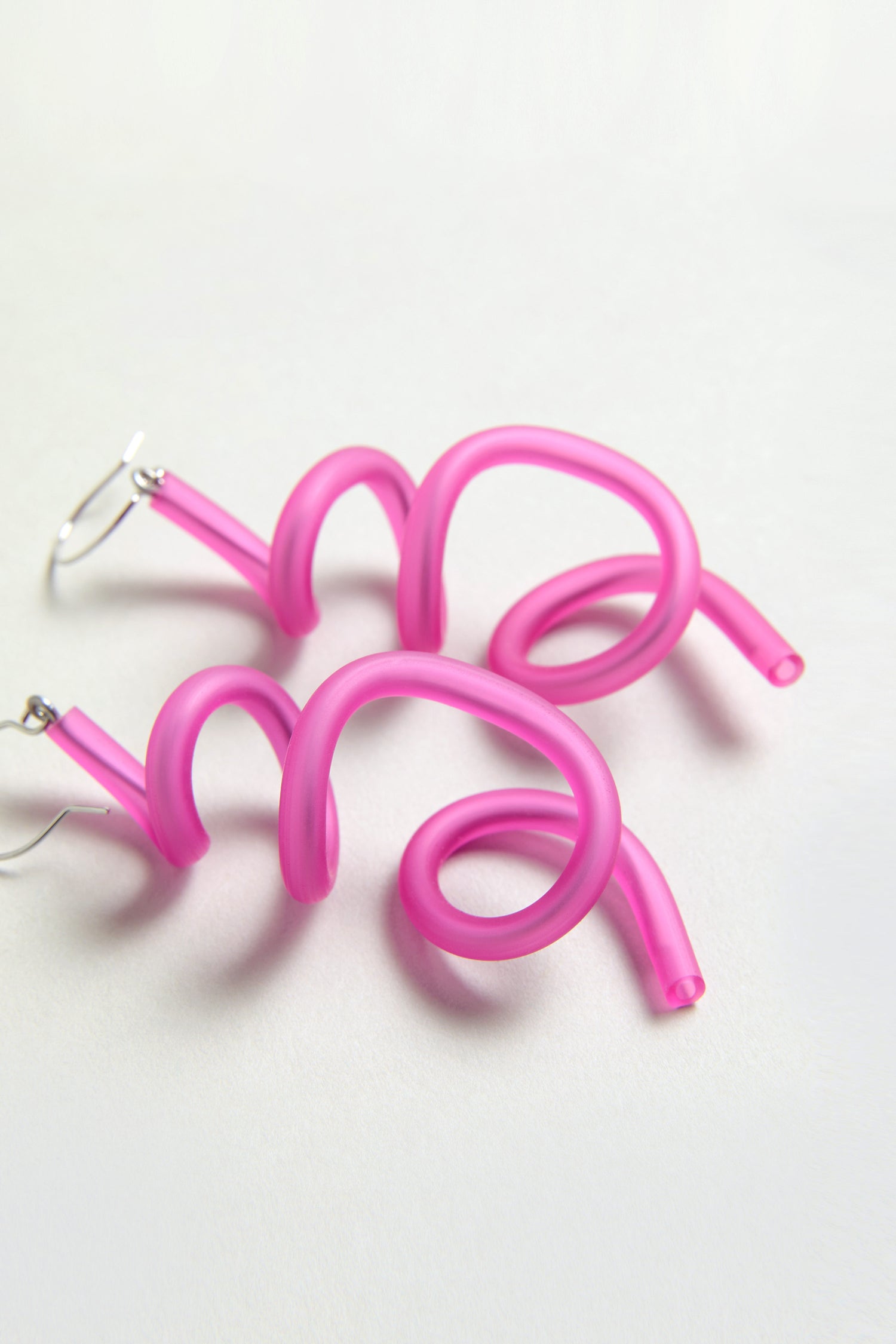 The Sketch Earrings showcase bright fuchsia, spiral-shaped tubes with silver hooks, elegantly arranged on a light surface.
