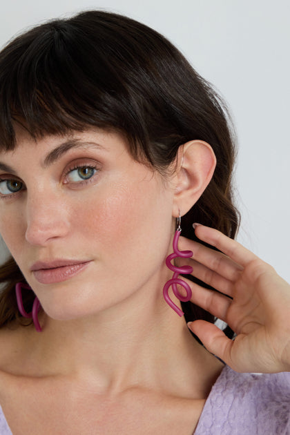 A woman with fair skin and brown hair touches her ear, showcasing the Sketch Earrings—large fuchsia sculptural statement pieces. She pairs them with a light purple textured top.