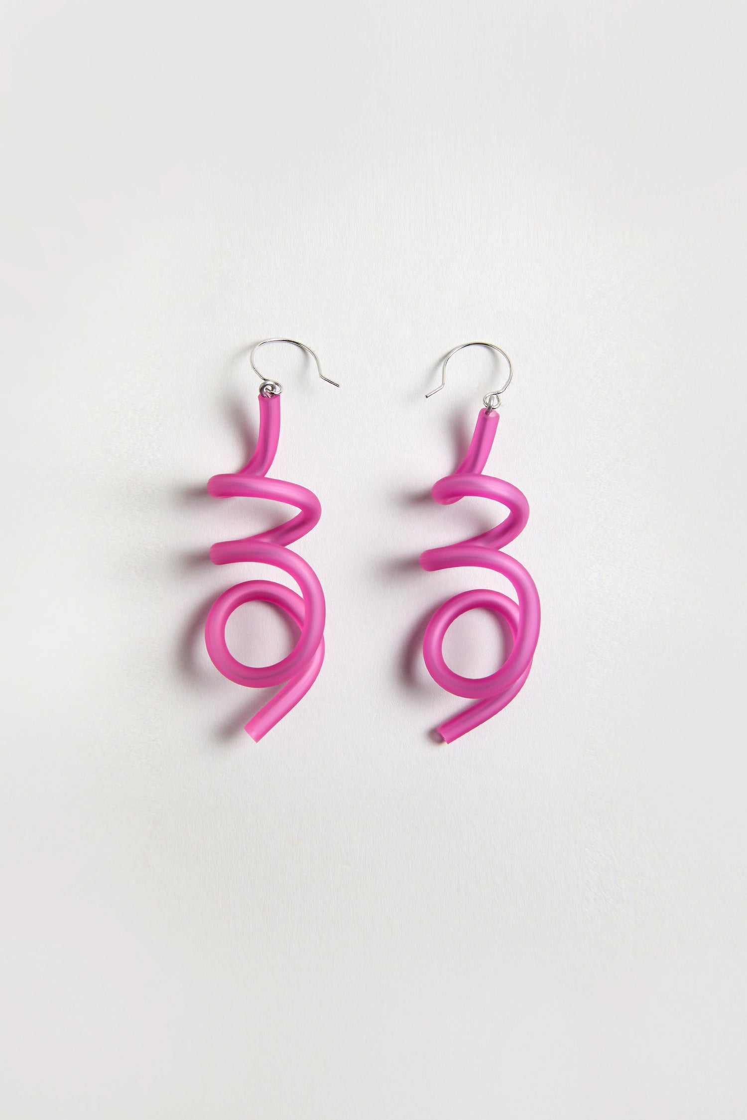 The Sketch Earrings feature bright fuchsia tubing twisted into abstract spiral shapes, creating a sculptural form. Displayed on a plain white background.